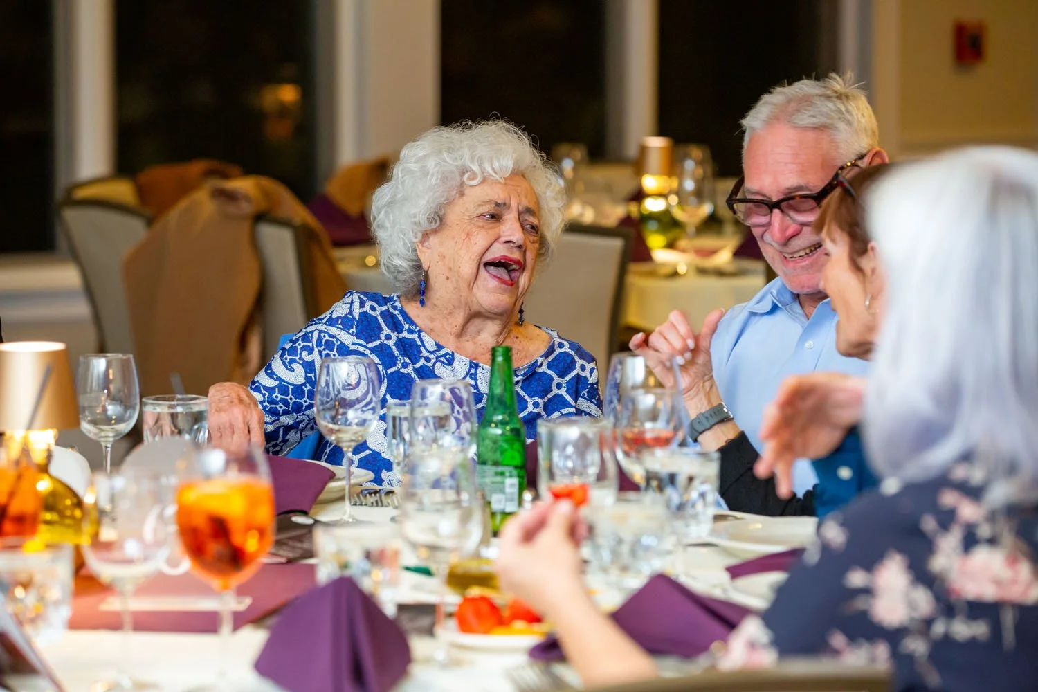 A group of elderly people laughing and talking at a dinner table in a restaurant or banquet hall.