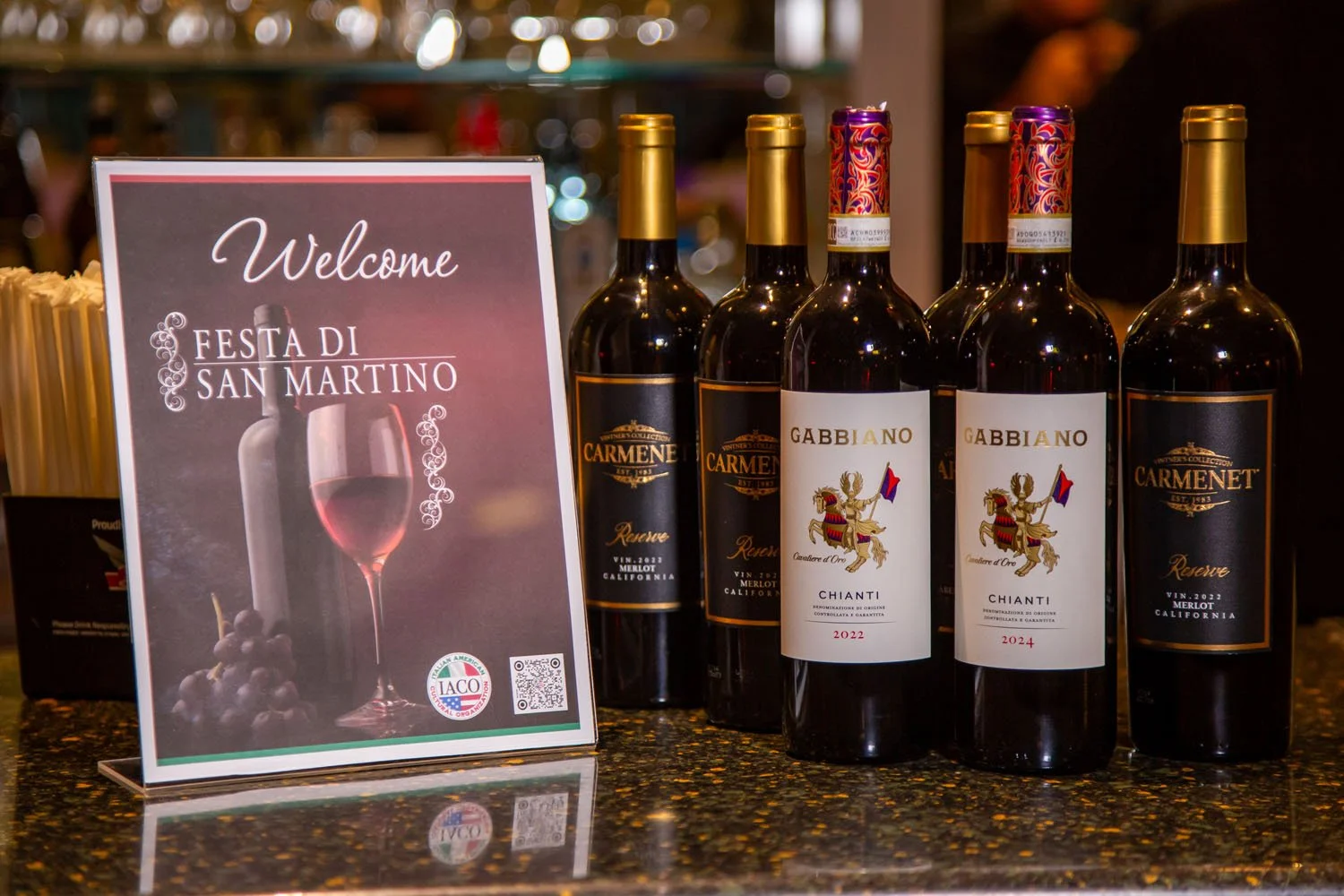 Wedding reception with bottles of wine and a welcome sign for Festa di San Martino, featuring a wine glass and grapes.