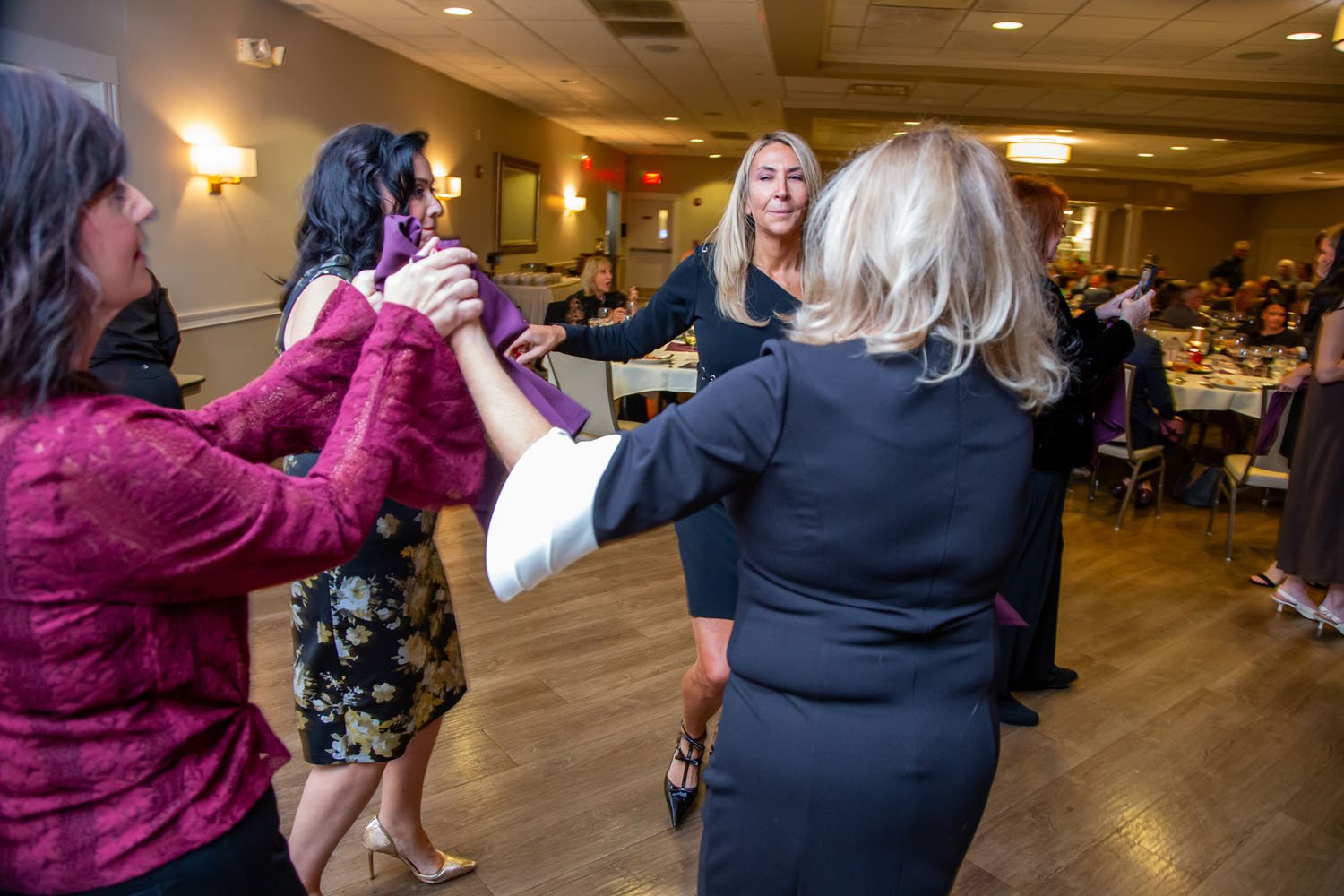 Four women are dancing and holding hands in a banquet hall with tables and seated guests in the background.