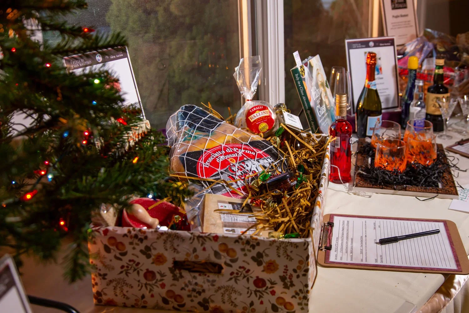 A Christmas gift basket on a table containing food items, surrounded by drinks and holiday decorations, next to a decorated Christmas tree with colorful lights.