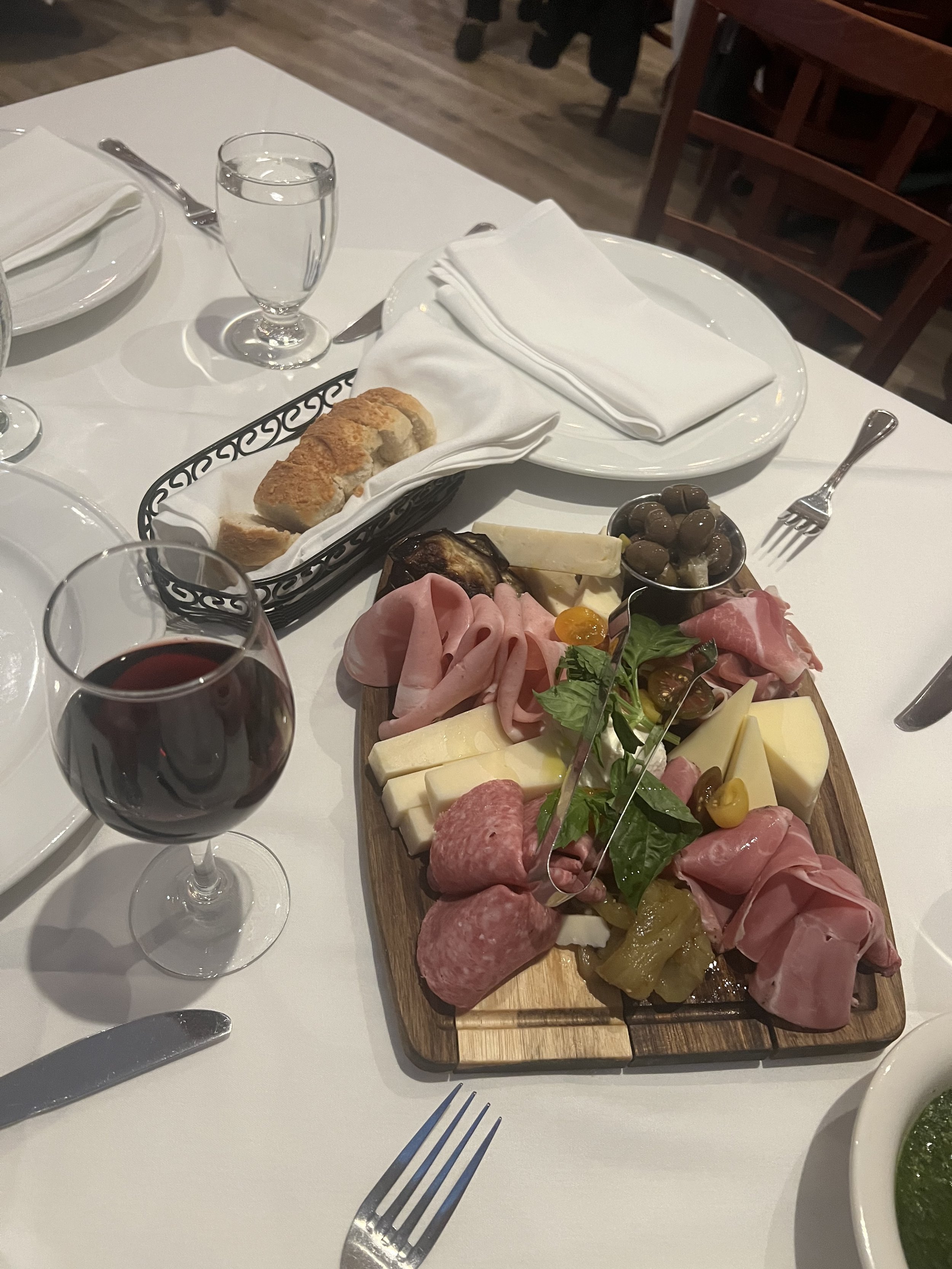 Dining table with charcuterie board featuring meats, cheeses, vegetables, a glass of red wine, and a basket of bread.