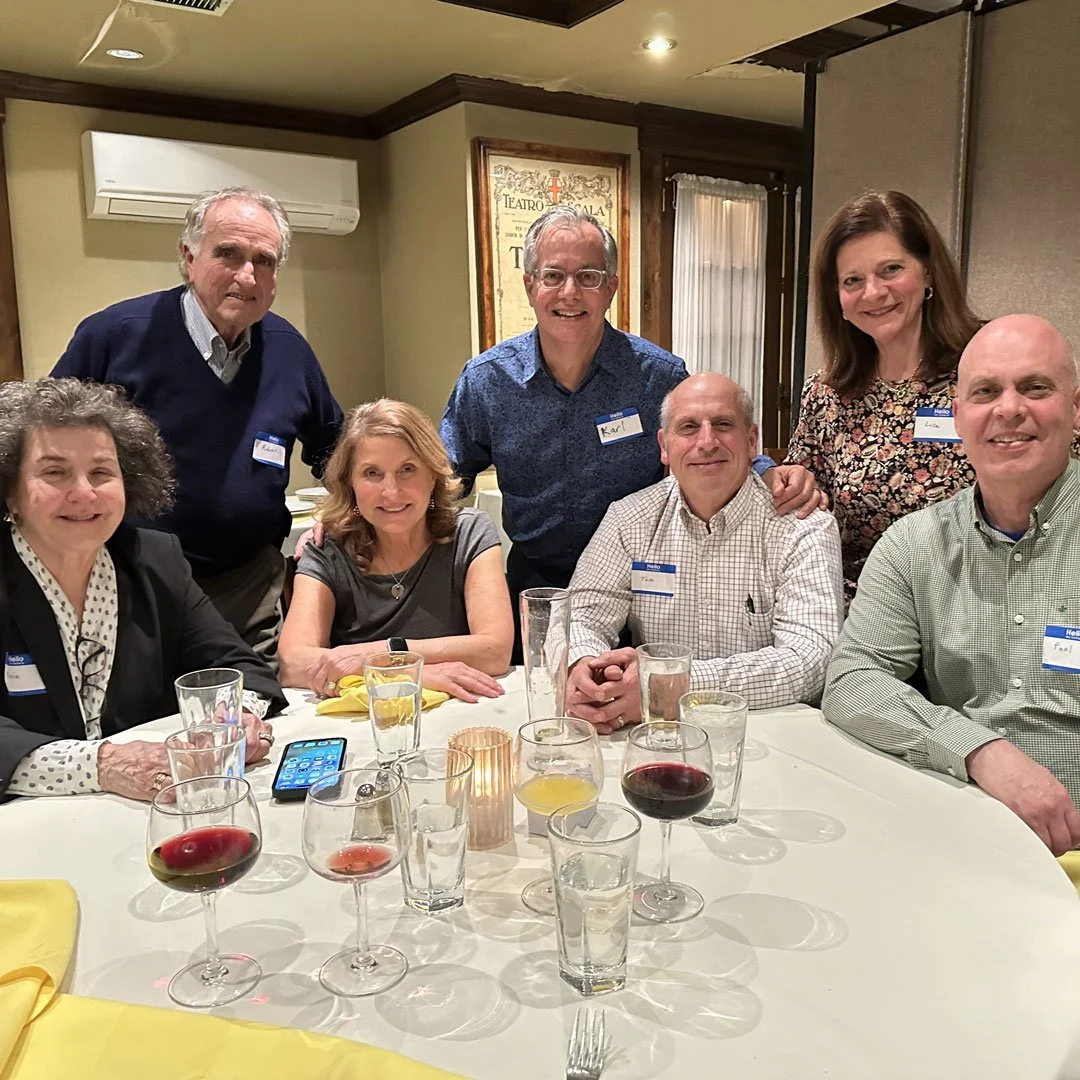 A group of seven people gathered around a table at a social event in a restaurant or banquet hall. They are smiling at the camera, with glasses of wine and water on the table. Four individuals are seated, and three are standing behind them.