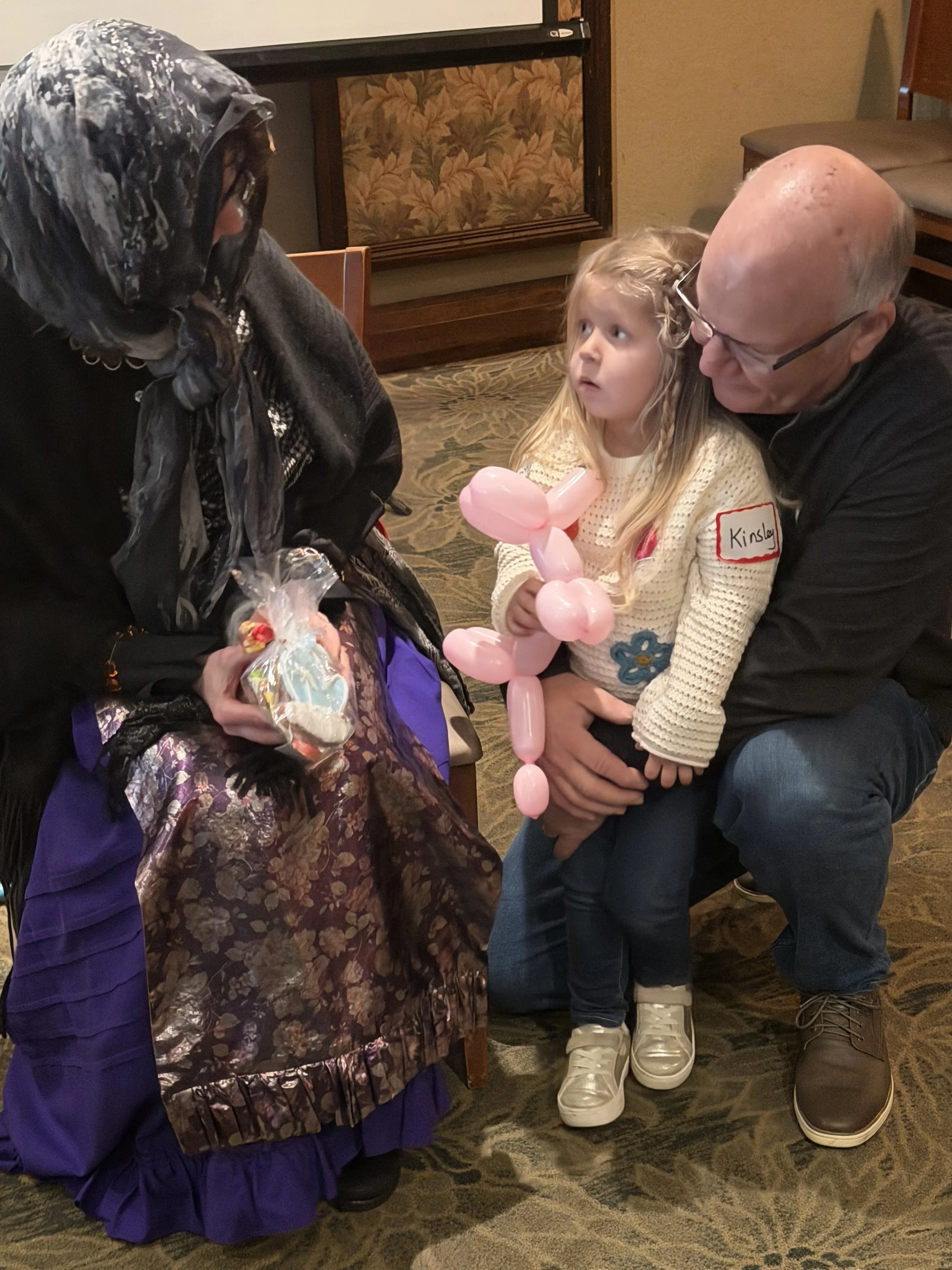 A girl with a pink balloon art face mask talking to a woman dressed in a witch costume and a man kneeling beside her. The girl has a name tag that says 'Kinsley' and appears to be at an indoor event.