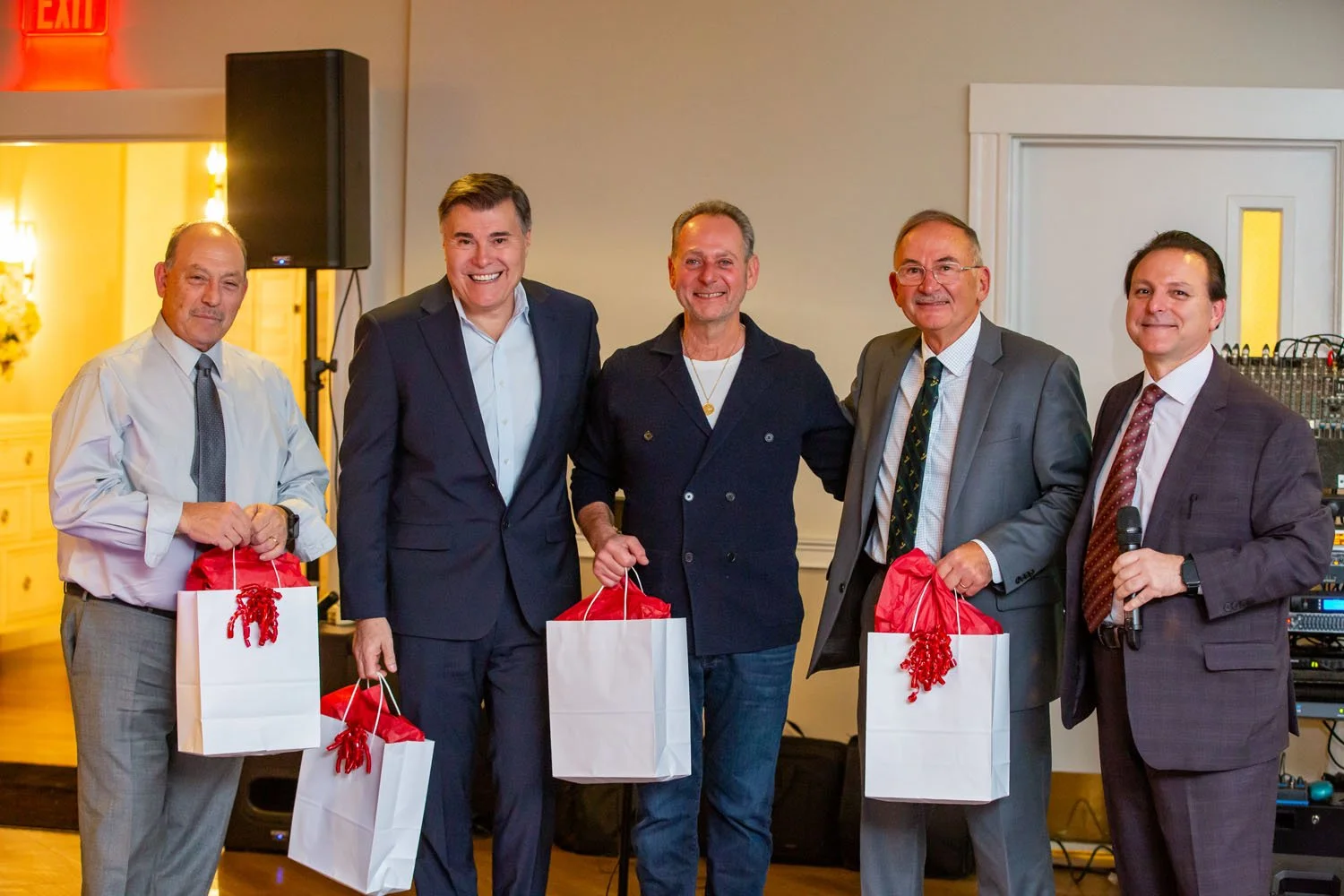 Group of five men standing indoors, each holding a white gift bag with red tissue paper, smiling for the camera at a celebration event.