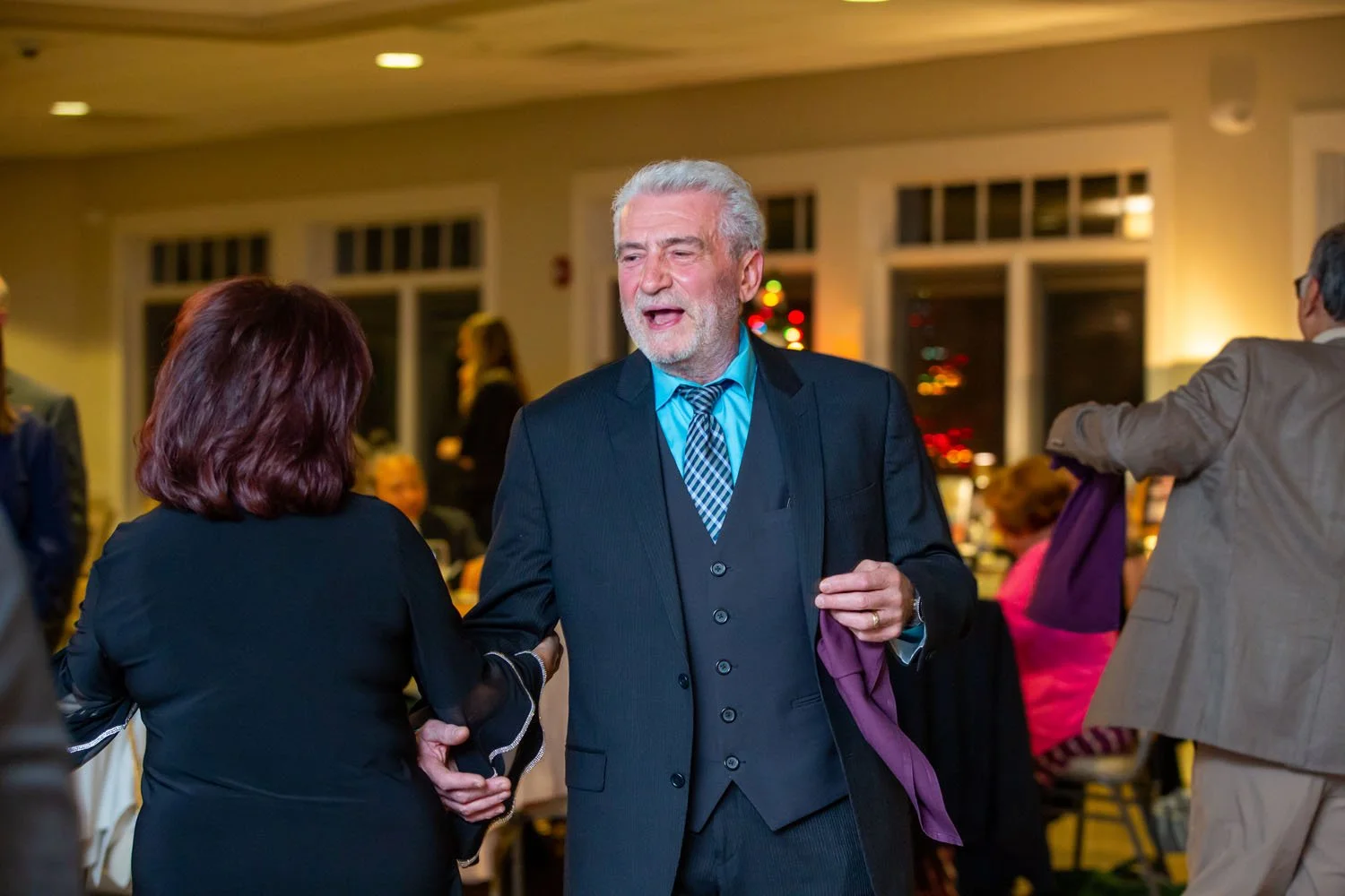 An elderly man with gray hair and beard in a dark suit and blue shirt, talking and shaking hands with a woman at a social gathering or party