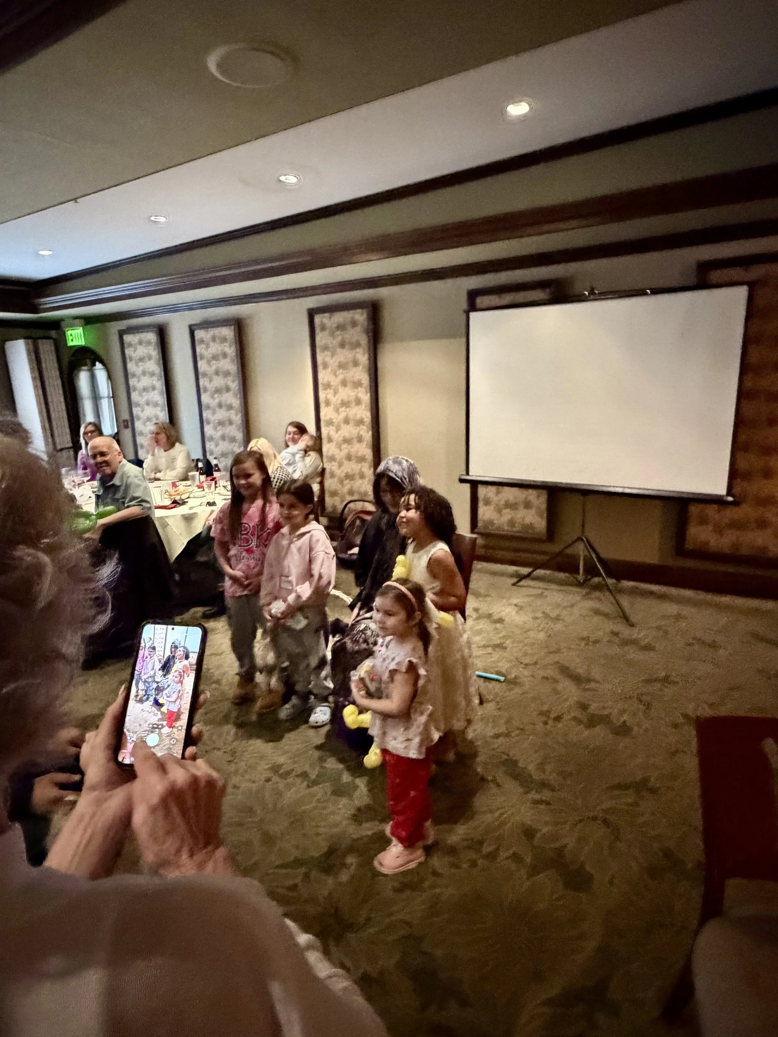 Children standing in front of a blank projection screen in a restaurant or banquet hall, with adults sitting at tables nearby, some taking photos.