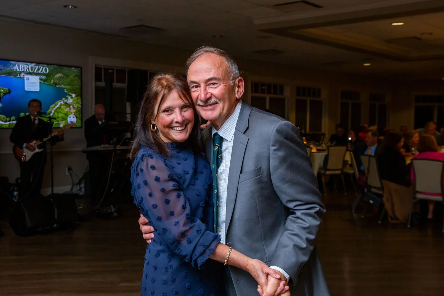 A smiling couple dancing at a formal event, with a musician in the background playing guitar and other guests seated at tables in a banquet hall.