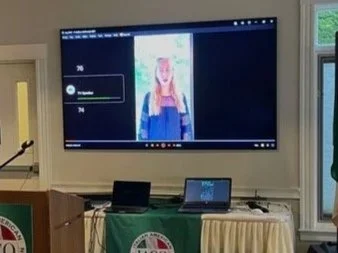 A woman is giving a presentation in a conference room with an American flag and an Italian flag. There is a large screen displaying a video call or presentation, and a table with laptops and a banner with the IACO logo.