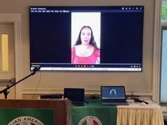 A presentation or speech setup in a room with American and Italian flags, a large screen displaying a woman, and a podium with the IACO logo.