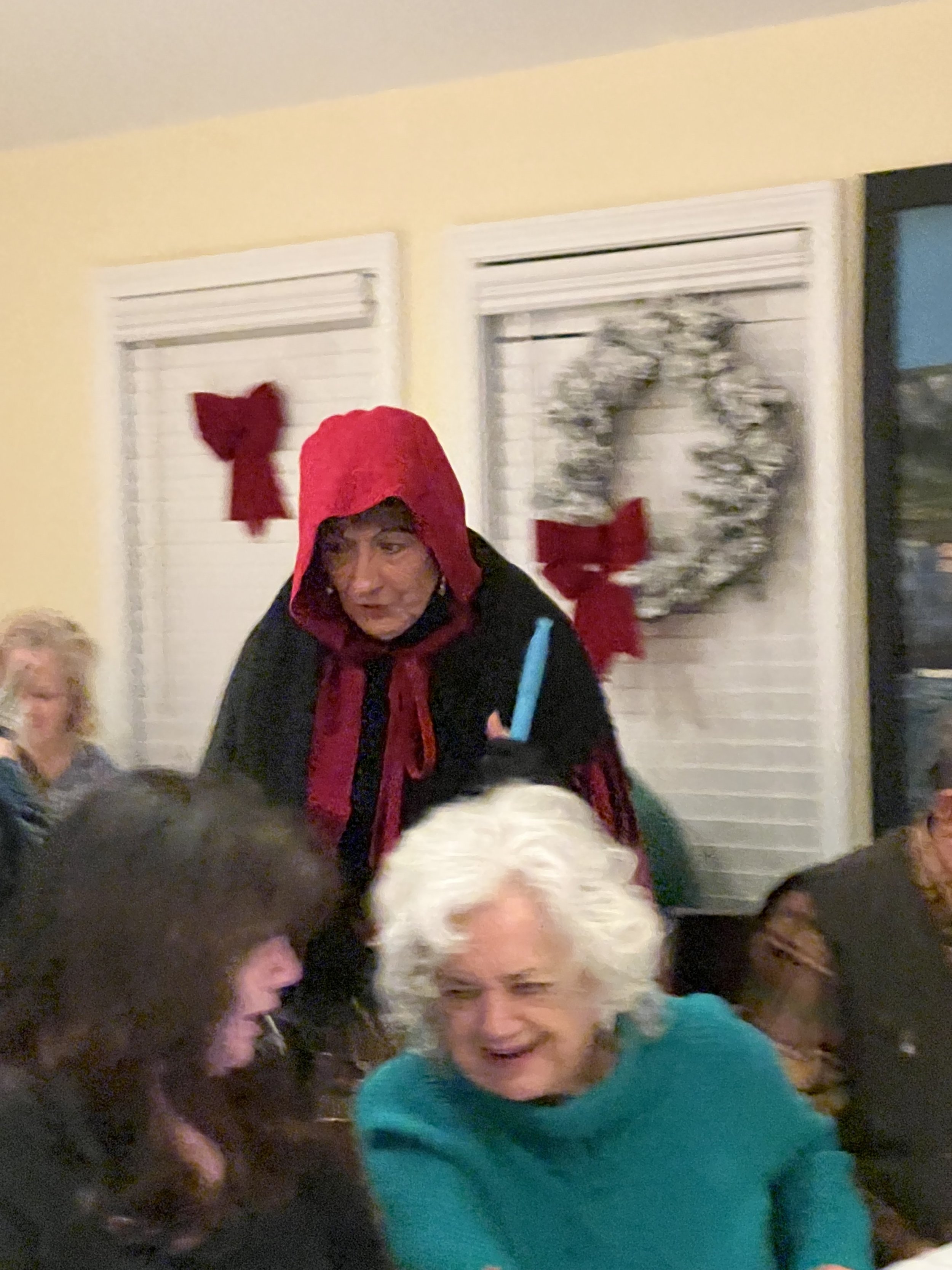 A person in a red hooded cloak stands in a decorated room with holiday wreaths and bows on the windows. In the foreground, two people are engaged in conversation, one with white hair wearing a teal sweater.