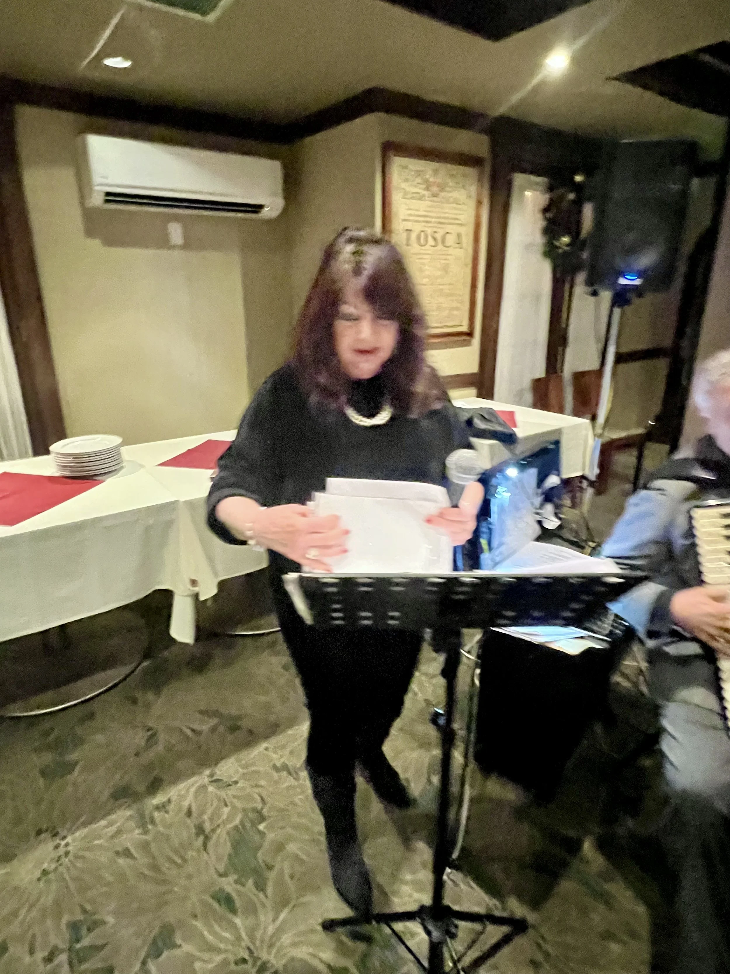 A woman in black clothing reading from papers at a music stand during a performance, with a man playing an accordion beside her, in a decorated indoor setting.