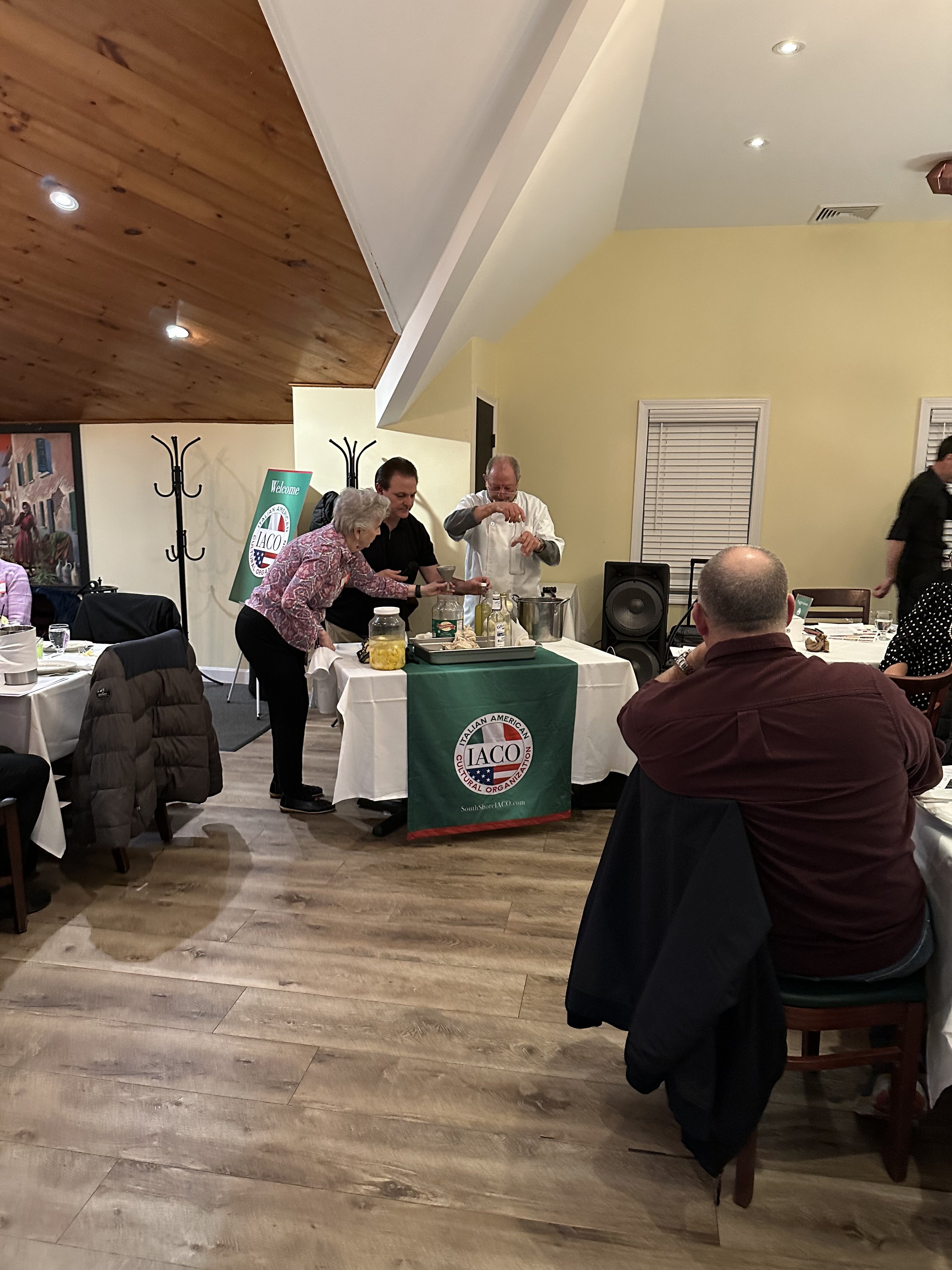 Three people preparing a dish at a table with ingredients and cooking equipment, under a sign for the Italian American Cultural Organization, inside a restaurant with wooden ceiling and several seated guests.