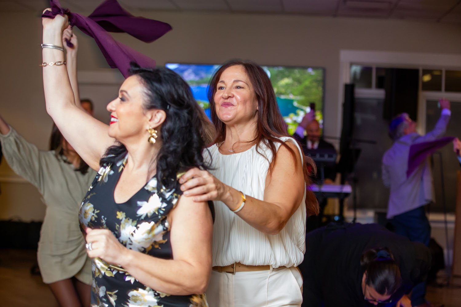 People dancing at a party or celebration, with a woman in the foreground holding a purple cloth and another woman behind her smiling.