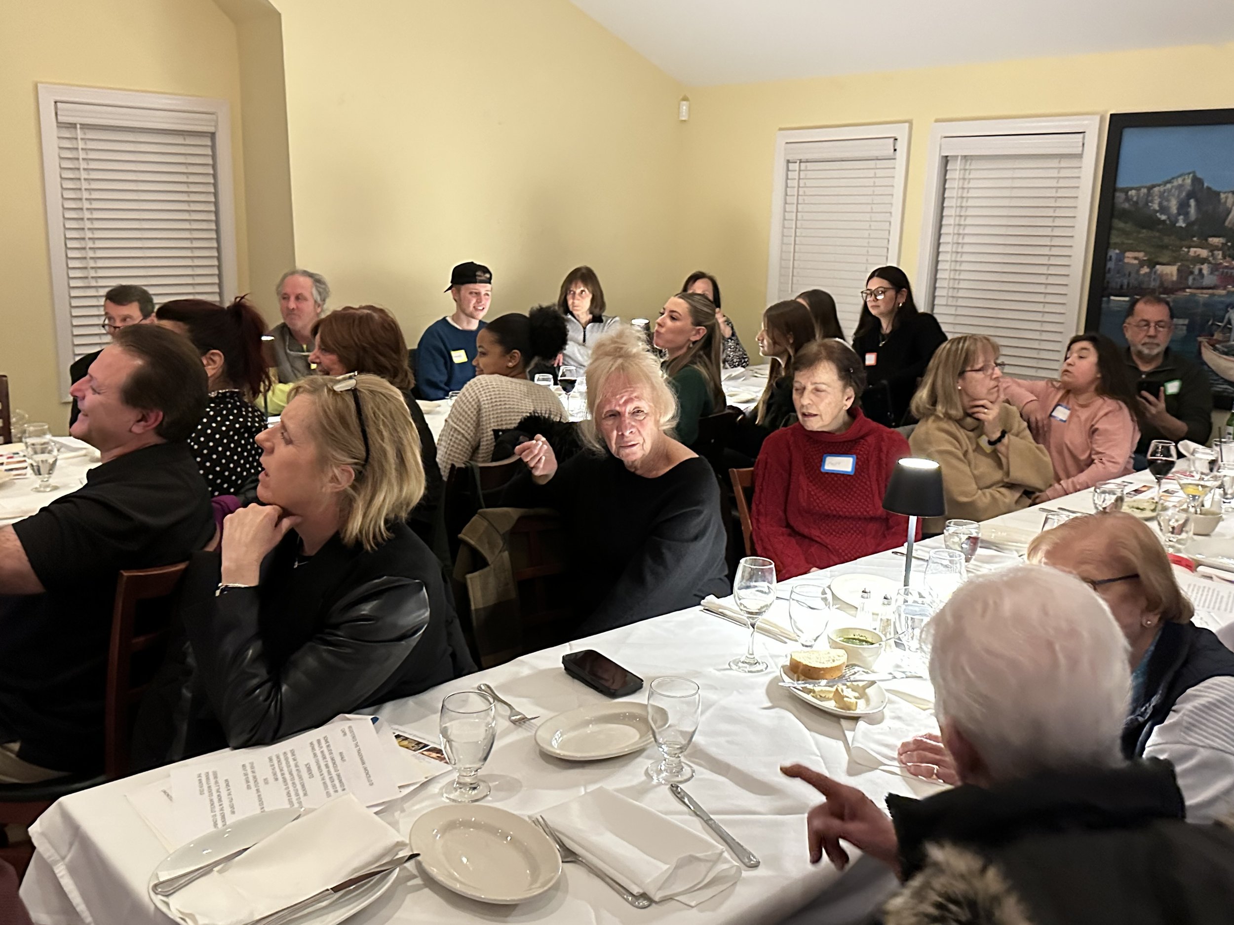 Group of people seated at a community event or gathering in a dining area, with white tablecloths, plates, and glassware on tables. They appear to be engaged in conversation or listening to a speaker.