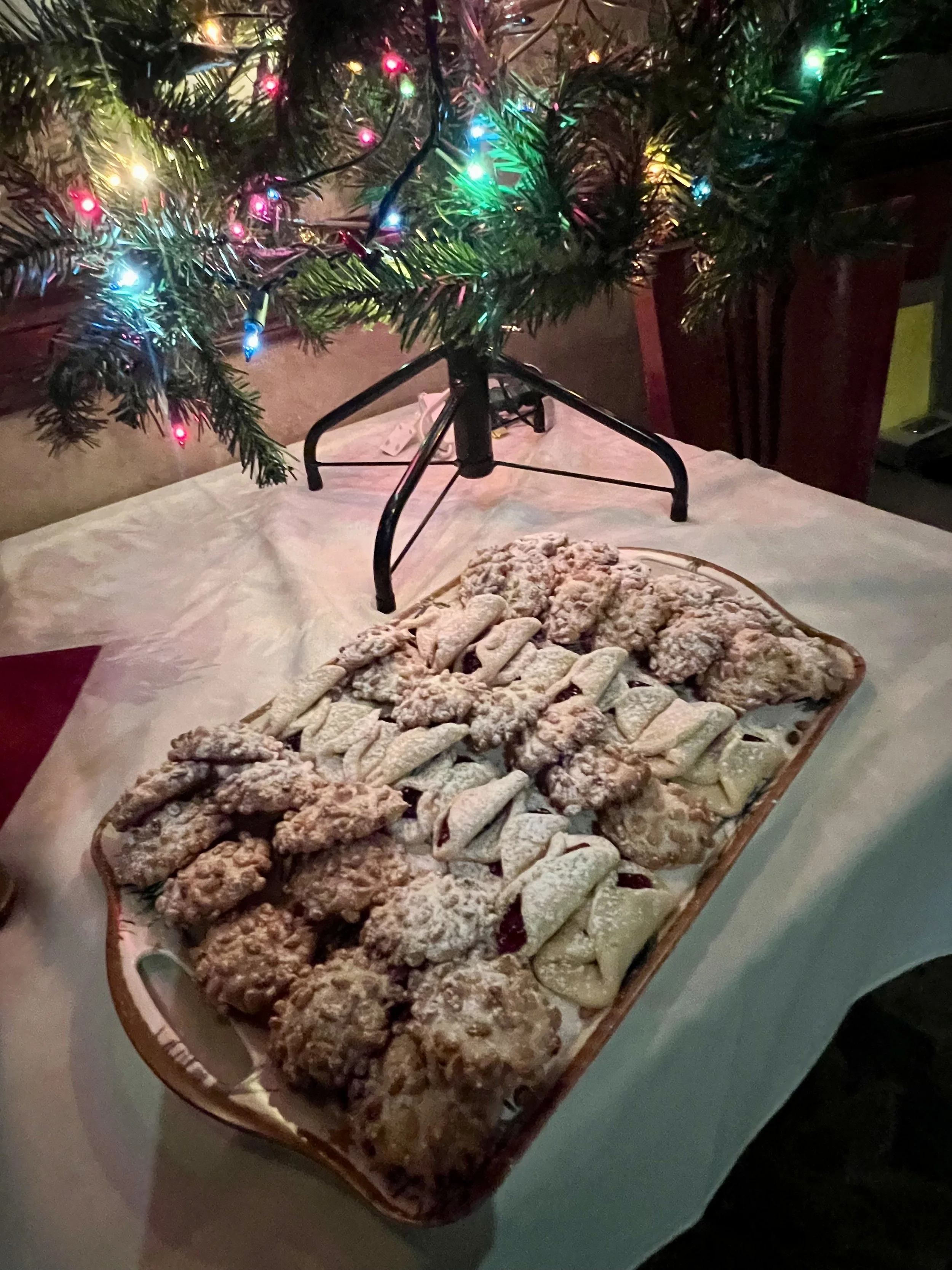 A tray of assorted Christmas cookies dusted with powdered sugar on a table decorated for Christmas, with a small Christmas tree with colorful lights in the background.