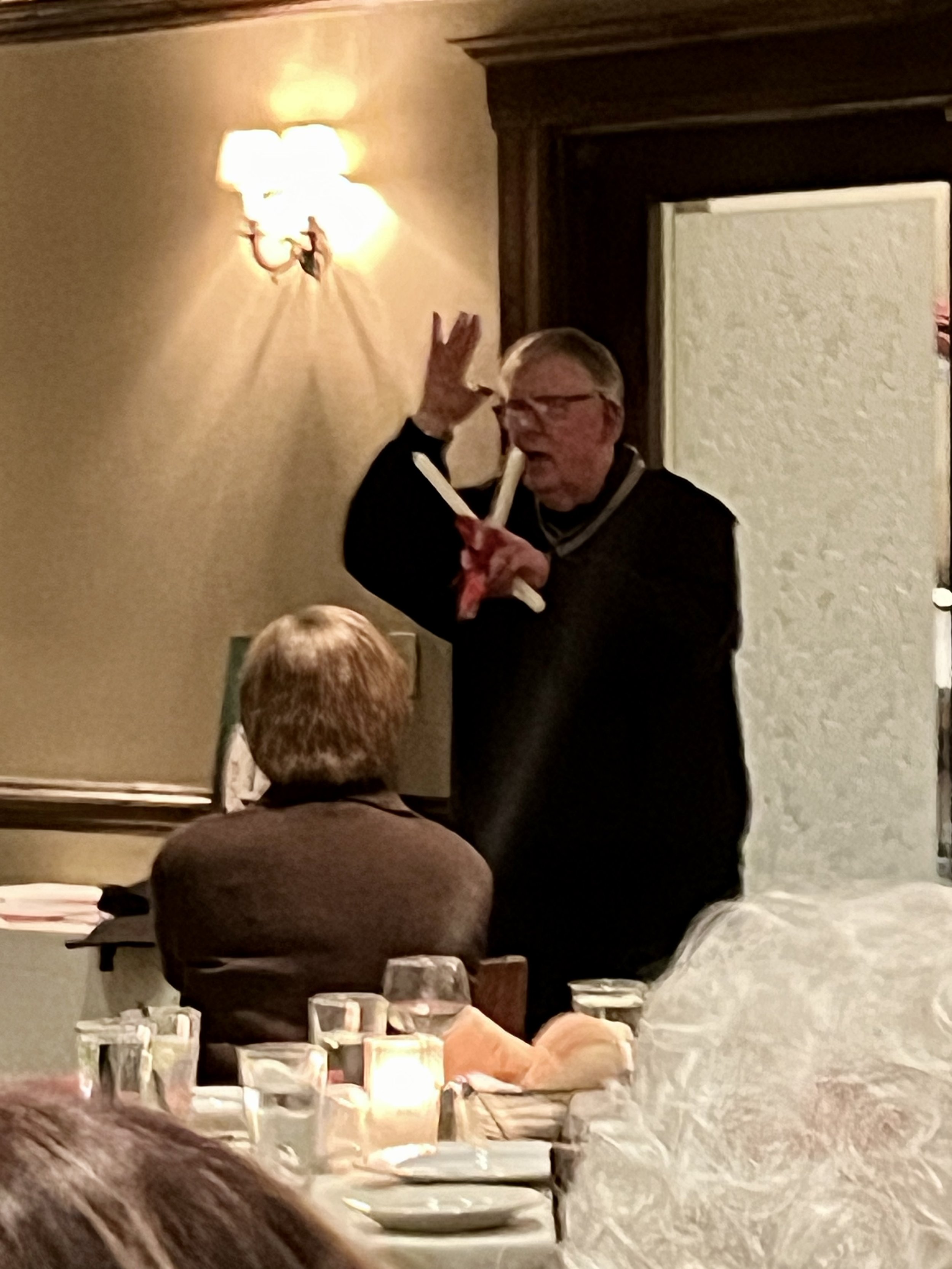 Older man speaking to audience in dimly lit room, holding a paper with red markings, near dining table with glassware and candles.