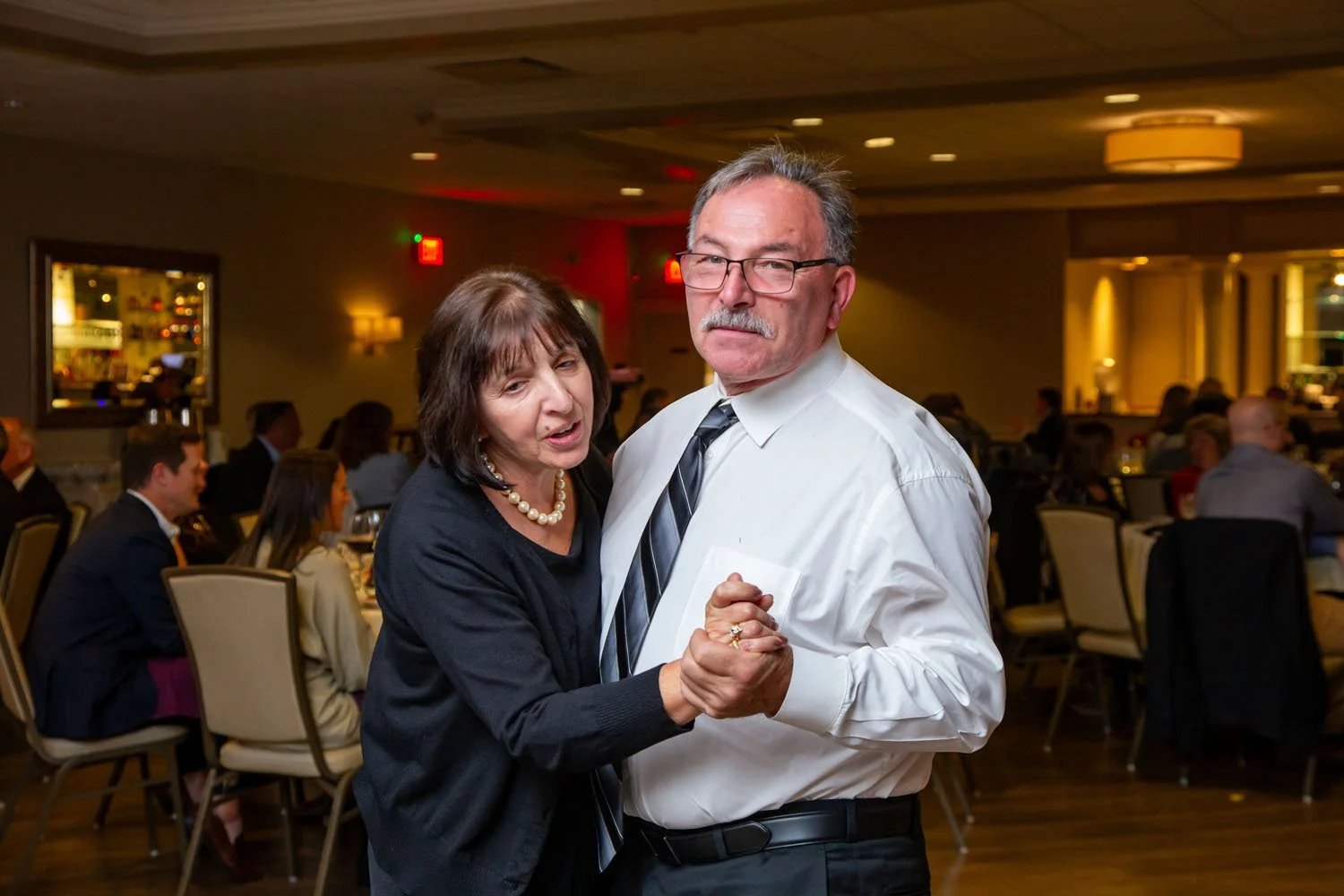 A senior couple dancing together at a formal event in a banquet hall, with other guests seated at tables in the background.