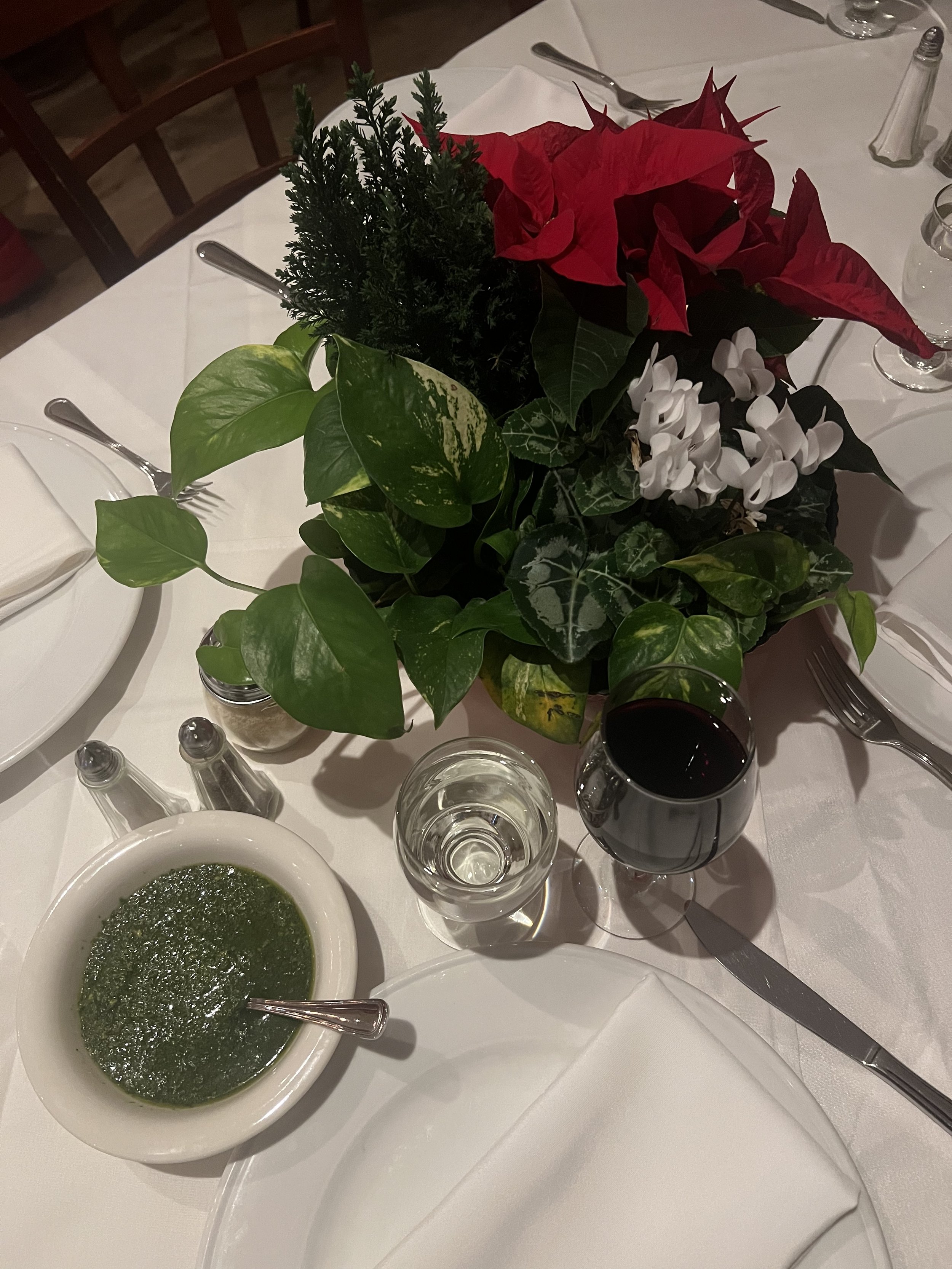 A dining table setup with a centerpiece of poinsettias, cyclamens, and green foliage. There's a bowl of green pesto-like sauce with a spoon, a glass of water, a glass of red wine, salt and pepper shakers, and a plate with a cloth napkin.