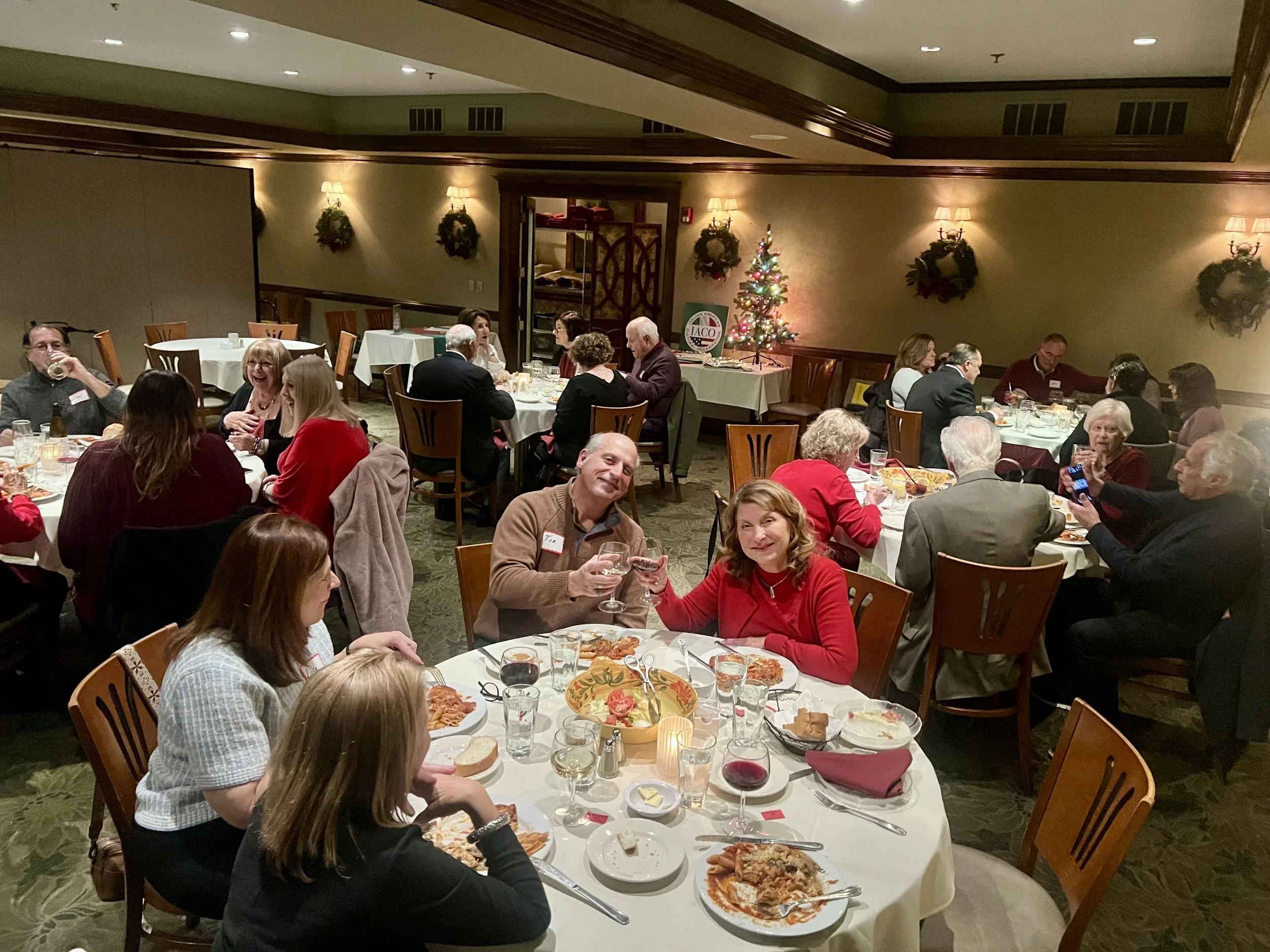People gathered at a Christmas dinner event in a decorated banquet hall with a Christmas tree and holiday wreaths, enjoying food and drinks.
