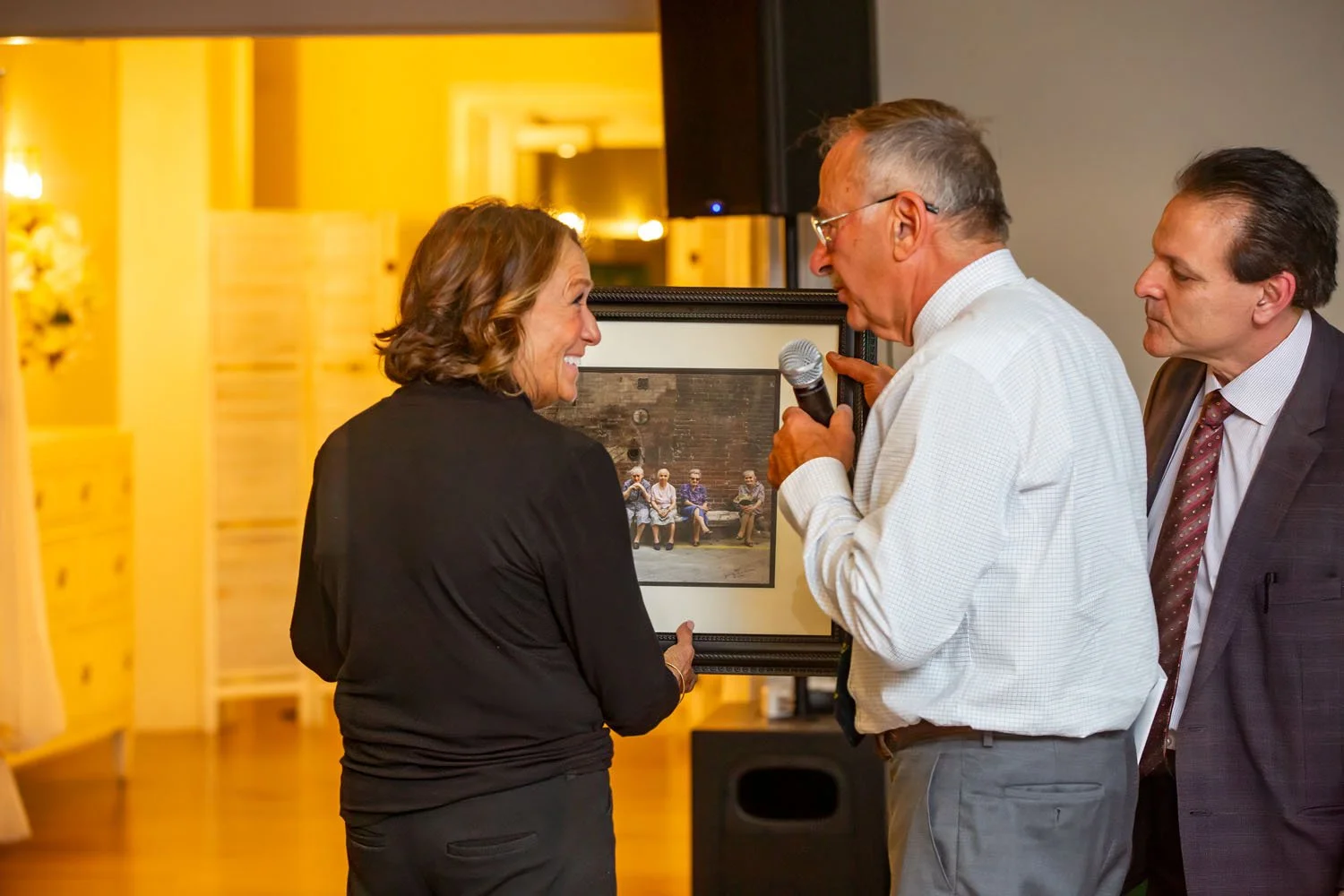 Three people at an art presentation, with a woman smiling and holding a framed photograph of a bench with four elderly people sitting on it. A man with glasses is speaking into a microphone, and another man is watching.