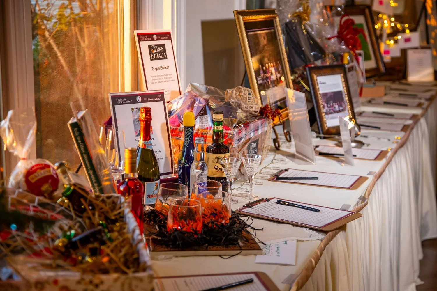 A table set up for a silent auction, featuring framed photographs, gift baskets, bottles of wine, glasses, and bid sheets with pens. The setting is decorated with holiday lights and festive ornaments.