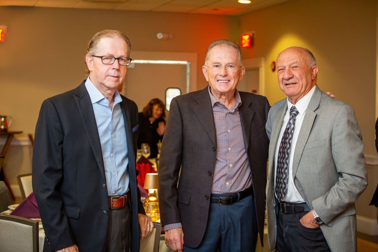 Three men in suits standing together at a formal event, smiling and posing for the photo with a woman in the background at a decorated banquet hall setting.