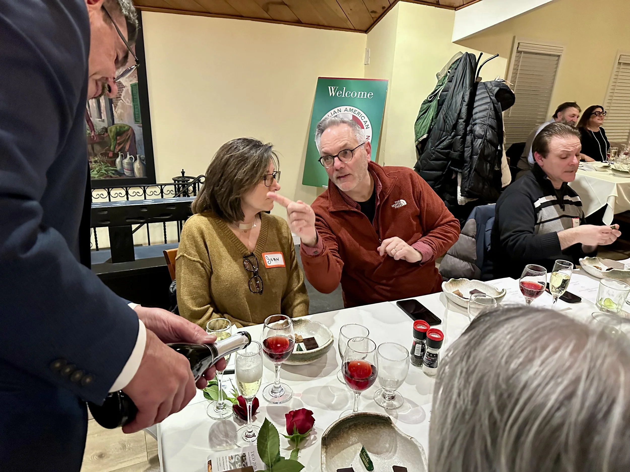 People gathered around a table at a restaurant or event, with drinks and appetizers, engaging in conversation. A man in a suit is pouring a drink, and a group, including a woman with glasses and a man with glasses and a red jacket, are talking. There