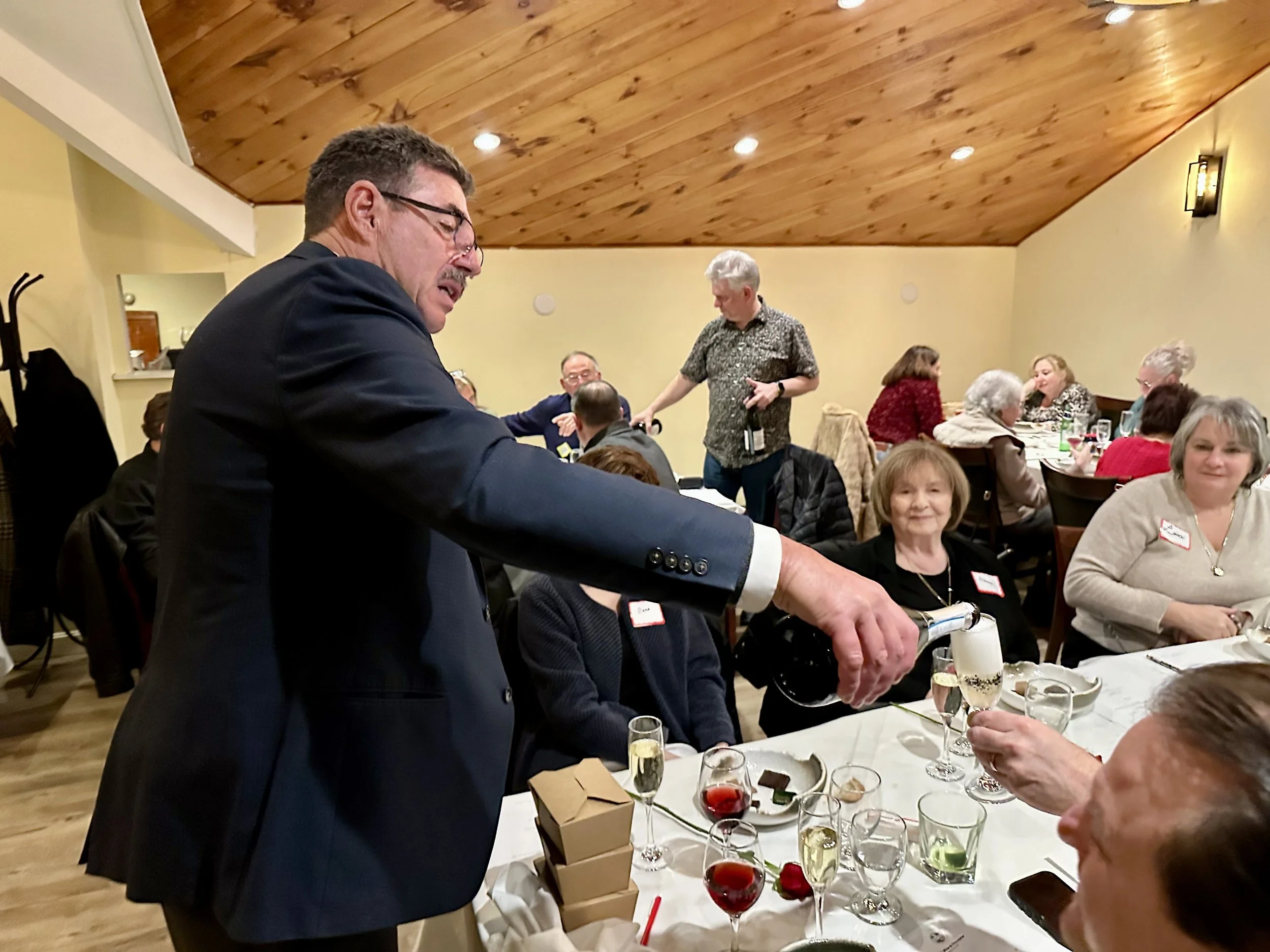 A man in a suit pouring champagne into a woman's glass at a social gathering, with several people seated at tables in a warmly lit room.