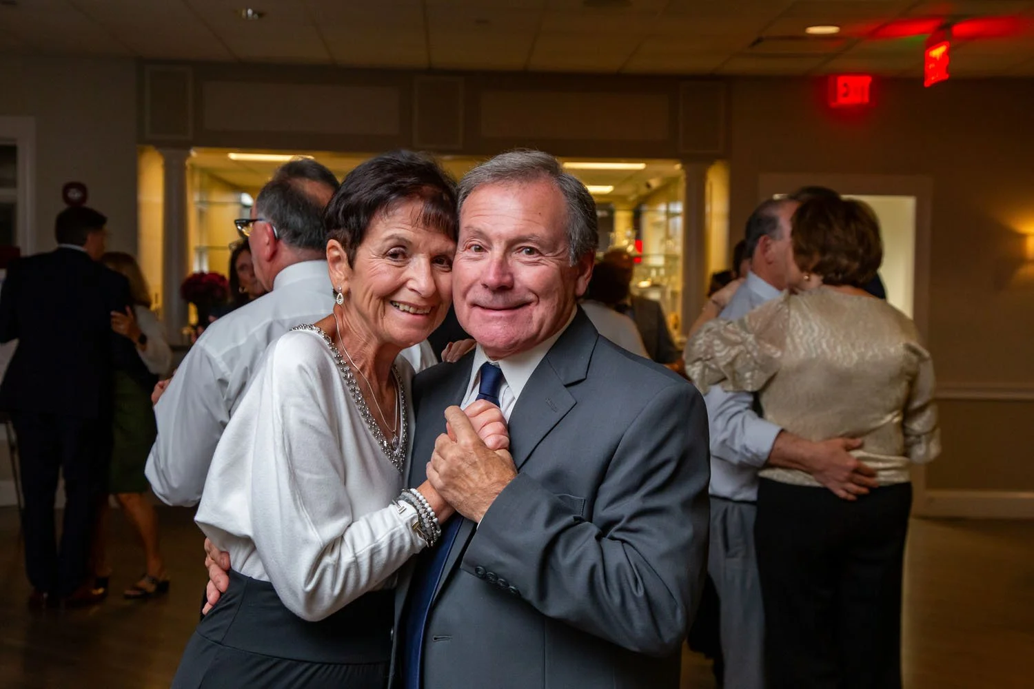 An elderly woman and man dancing and smiling at a social event, with other couples dancing in the background.