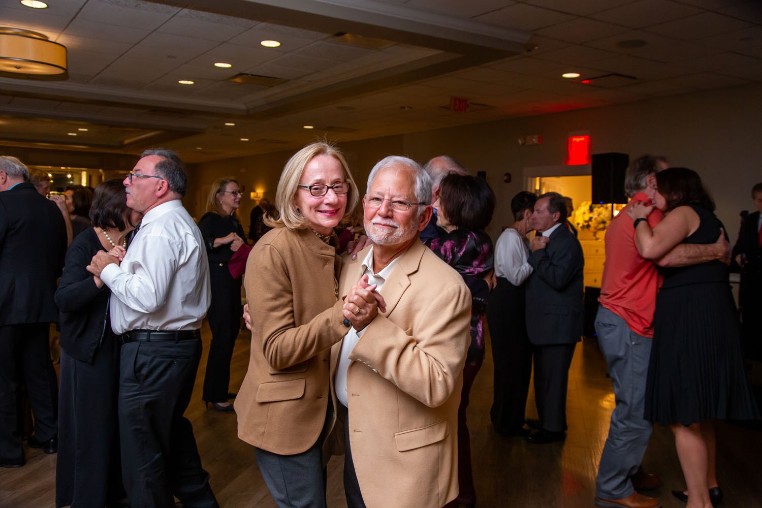 Older couple dancing closely at a social event with other couples in the background.