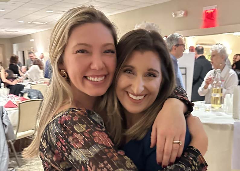 Two women smiling and embracing at a social event with people in the background.