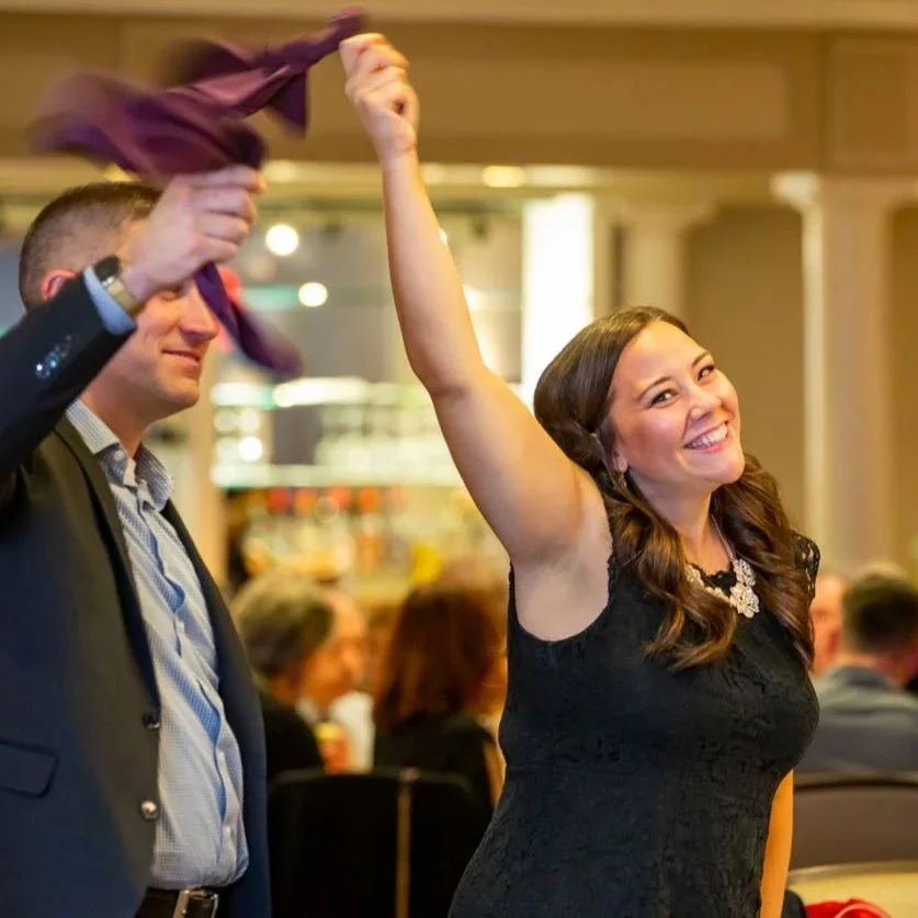 A woman in a black dress smiling and raising her arm while a man next to her, in a suit, is holding up a purple item, possibly a shirt or scarf, in a lively indoor setting with other people in the background.
