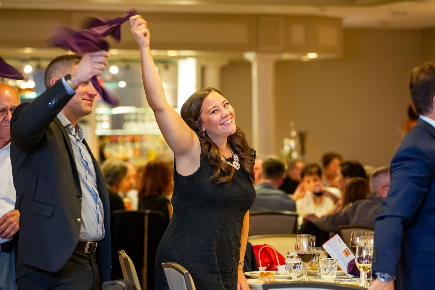 A woman in a black dress smiling and raising her hand at a banquet or formal event