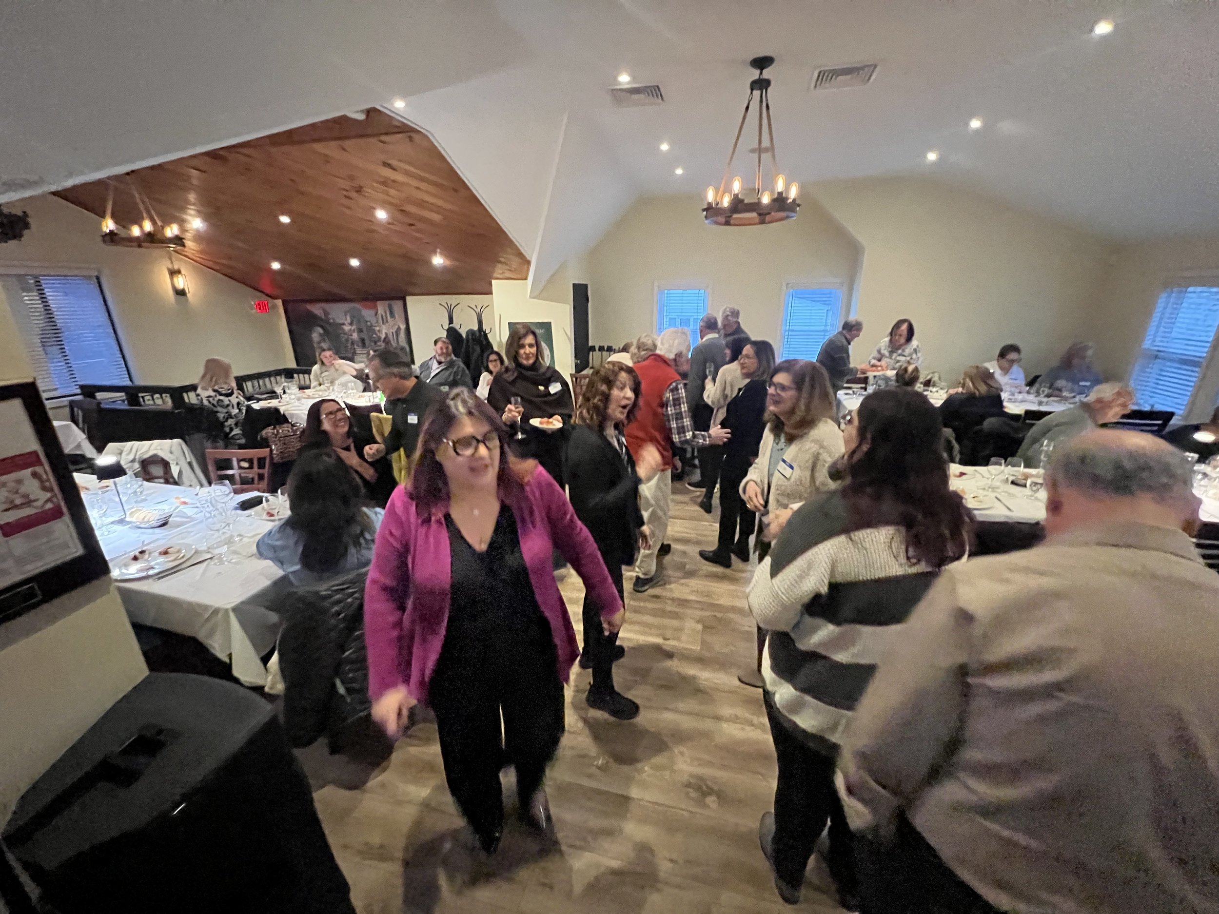 People gathered in a restaurant for a social event, wearing name tags, talking and dining. The room has wood paneling on the ceiling and a chandelier.