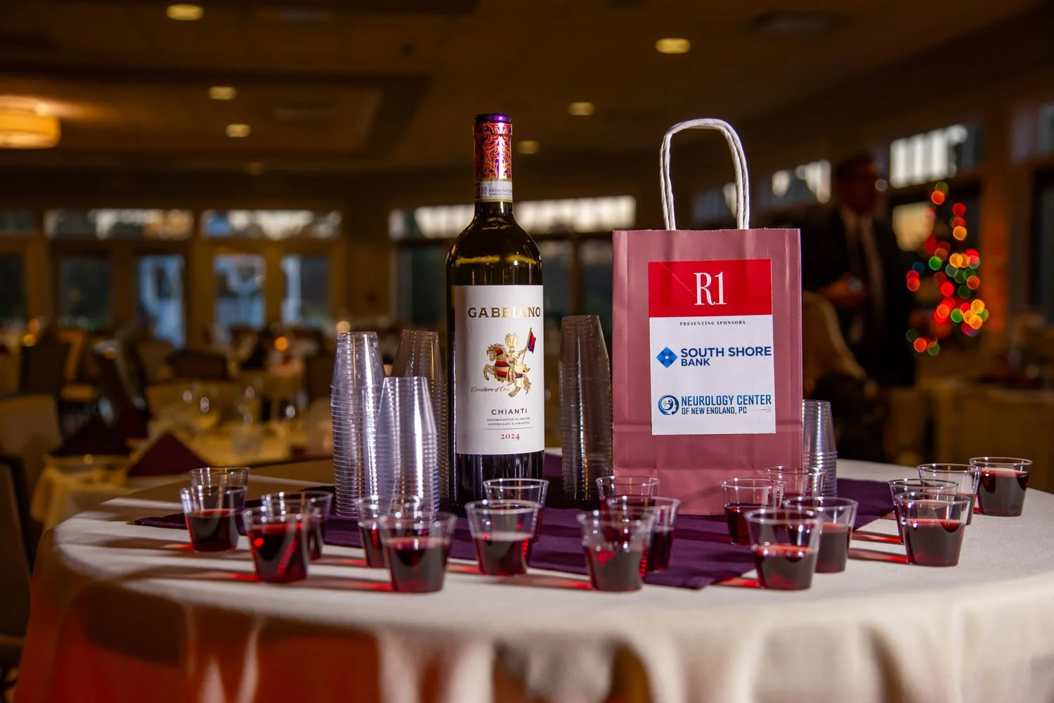 A round table with a large bottle of Gabelano Chianti wine, a pink gift bag with sponsors' logos, multiple small glasses filled with red wine, and clear plastic cups, set in a dimly lit banquet hall with a decorated Christmas tree in the background.