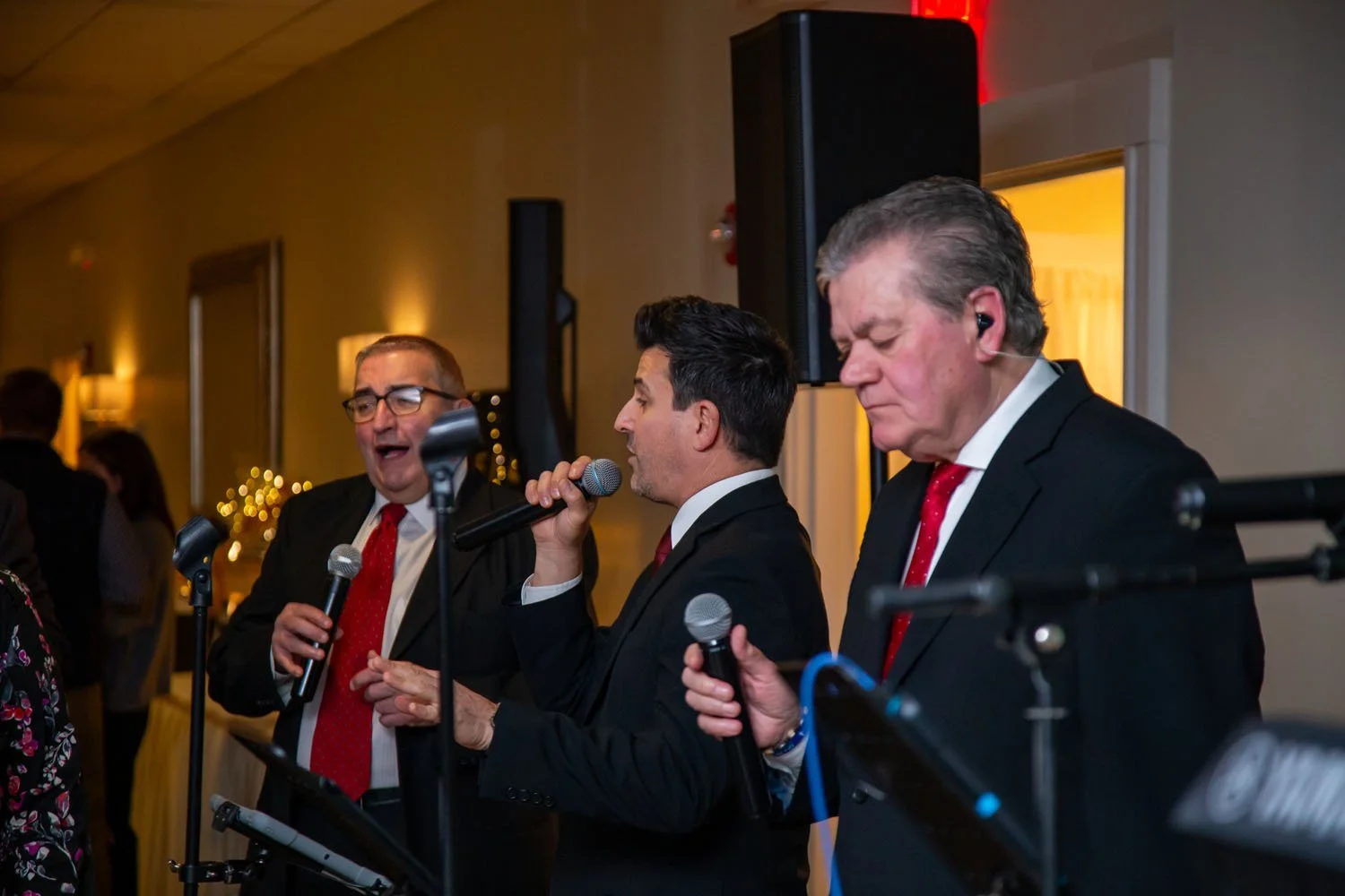 Three men in suits singing at a party, each holding a microphone, with one man wearing glasses and the other two with ear monitors.