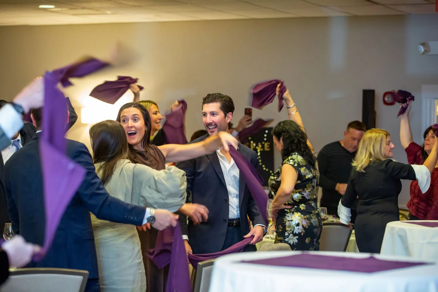 People celebrating at a formal gathering, throwing purple napkins in the air, with smiling and cheering faces.