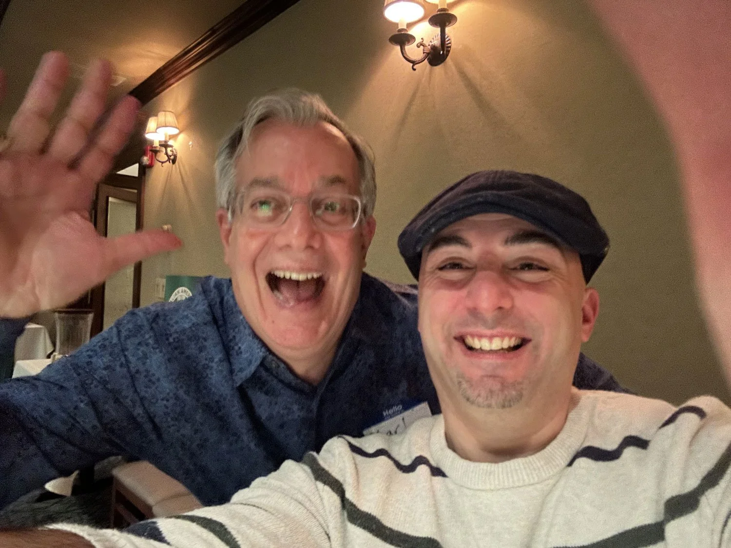 Two men taking a selfie at a restaurant, smiling and waving, with warm lighting and wall-mounted lamps in the background.