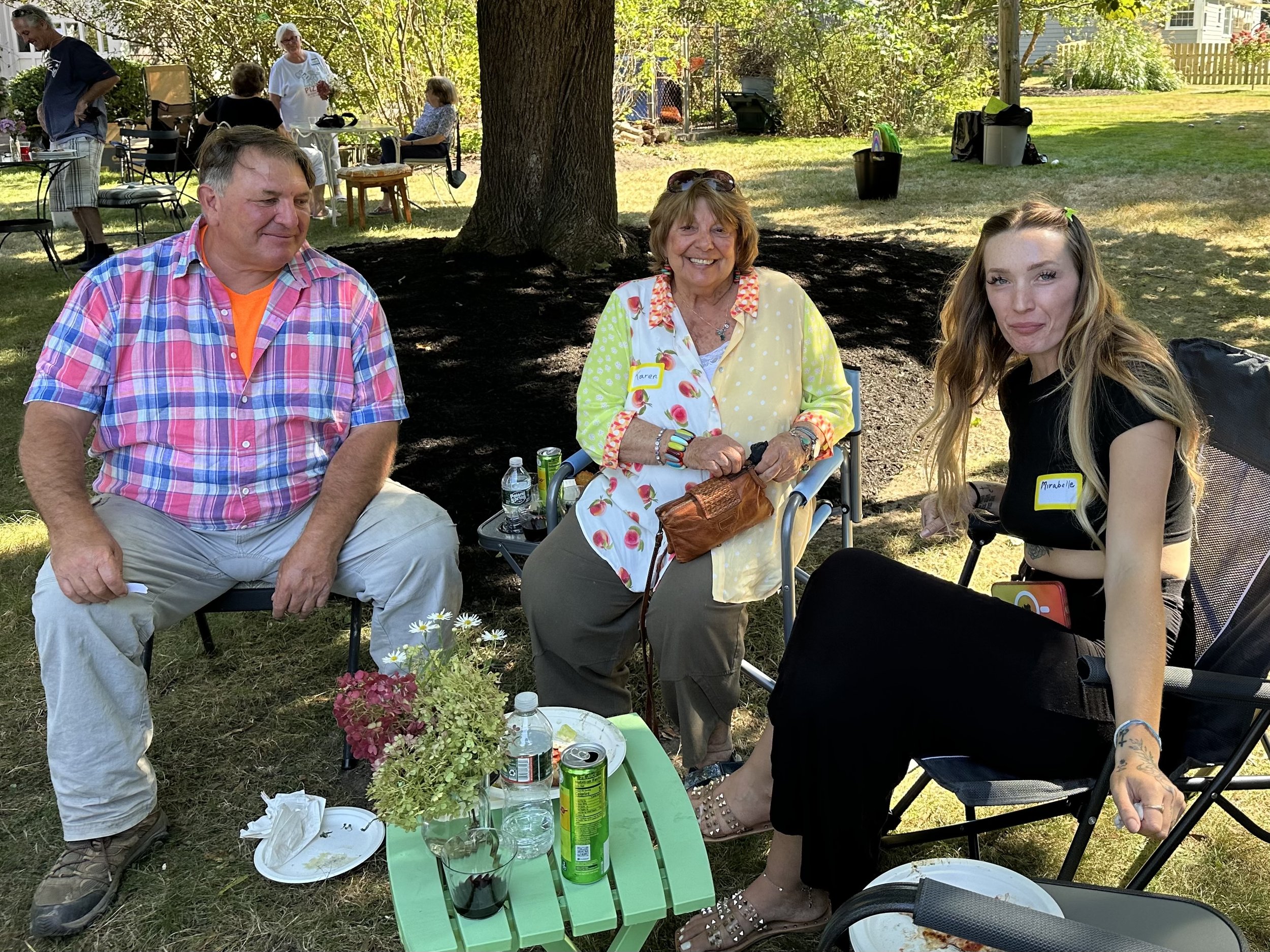 Three people sitting outdoors on chairs, at a gathering or picnic. The setting is a grassy area with trees and other people in the background. There are drinks and a small table with flowers in front of them.
