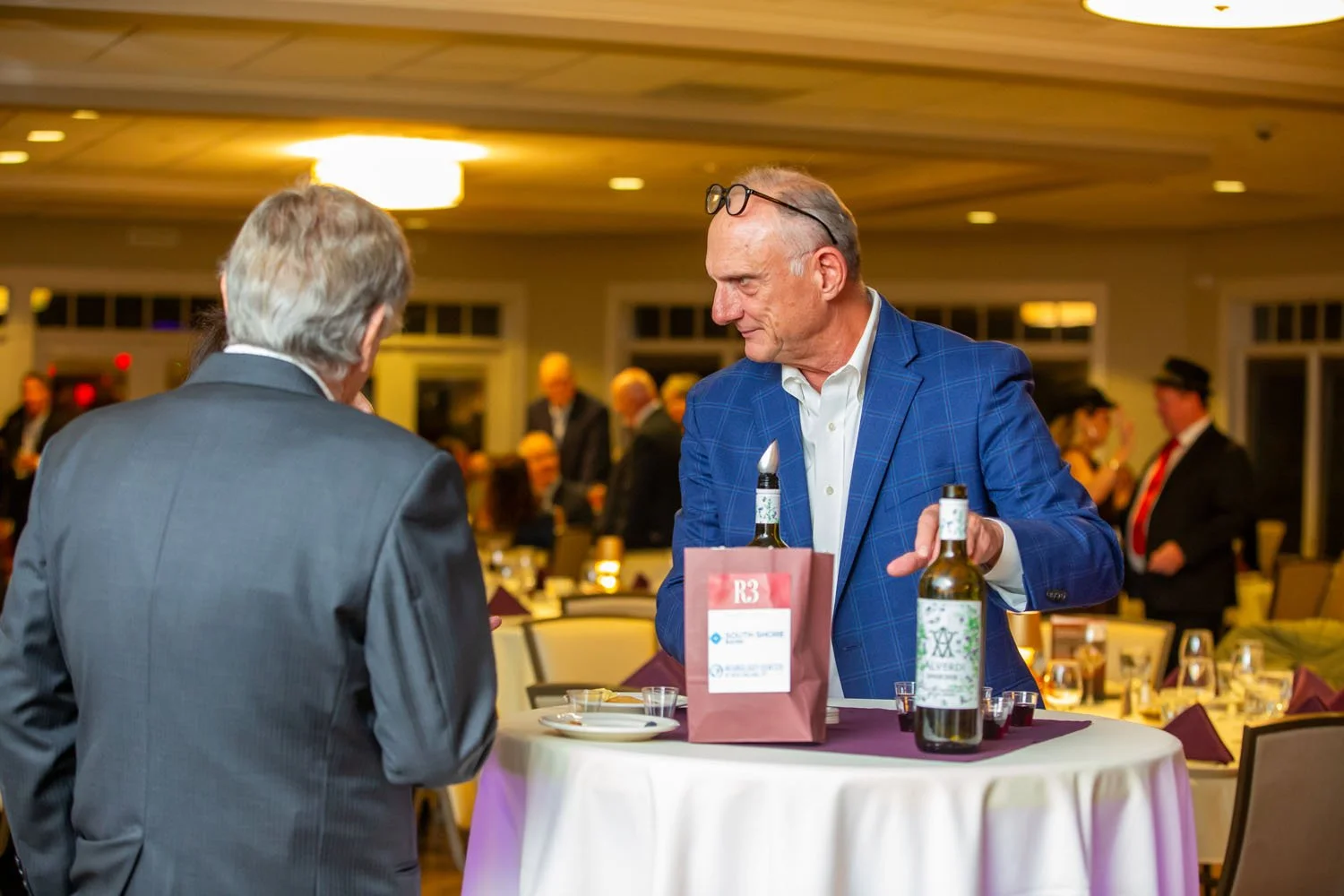 Two men in suits conversing at a social event with tables and other guests in the background.