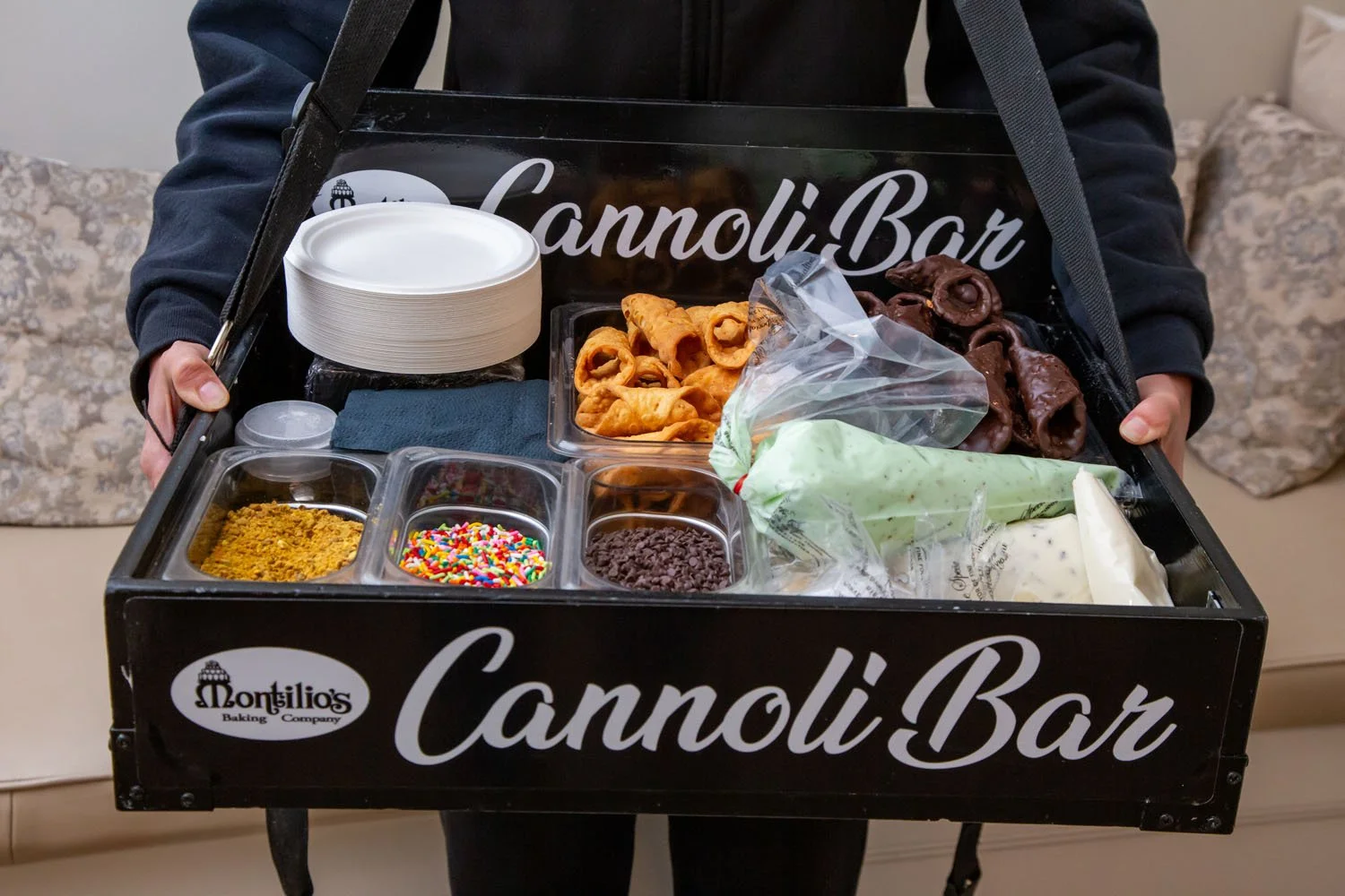 A person holding a black CanvasBar box filled with various toppings and ingredients for making cannoli, including fried shells, ricotta cheese, colorful sprinkles, chocolate chips, crushed cookies, and other toppings, with a stack of white paper plat