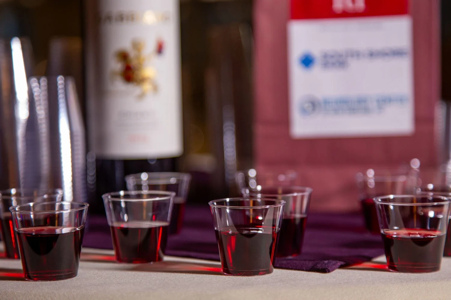 Six plastic cups filled with red wine arranged on a table, with a blurred background of bottles and a sign.