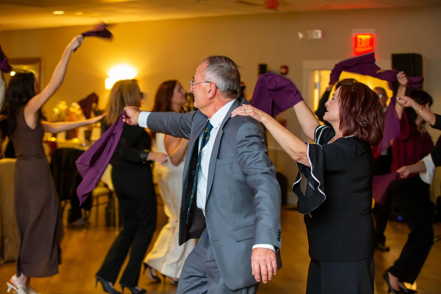 People dancing at a celebration event, with a man in a gray suit and a woman in black sharing a dance, surrounded by others holding purple cloths or scarves and dancing.