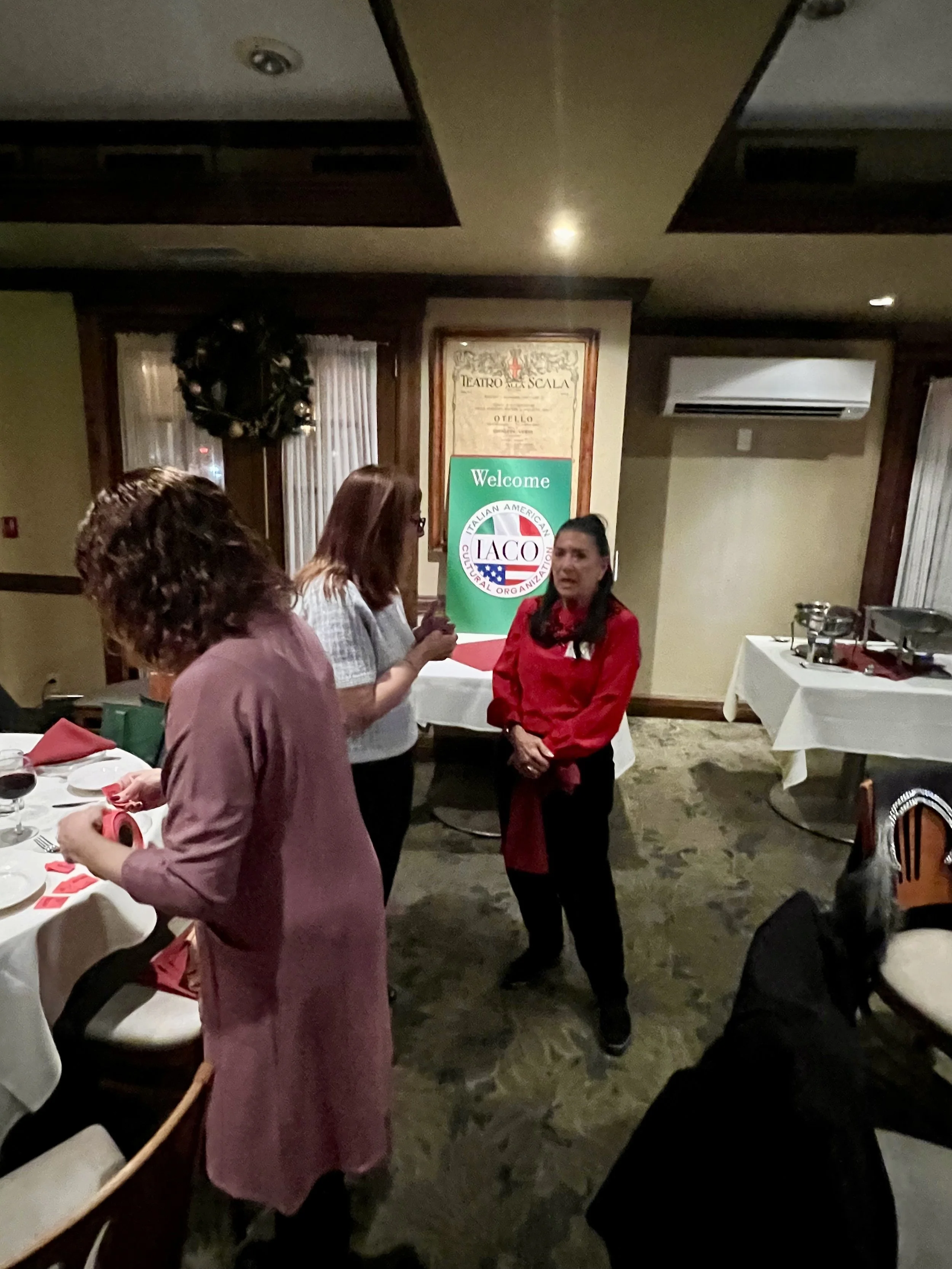 Three women are standing and talking near a table set for a meal in a restaurant, with a sign that says 'Welcome Italian American Cultural Organization' in the background.