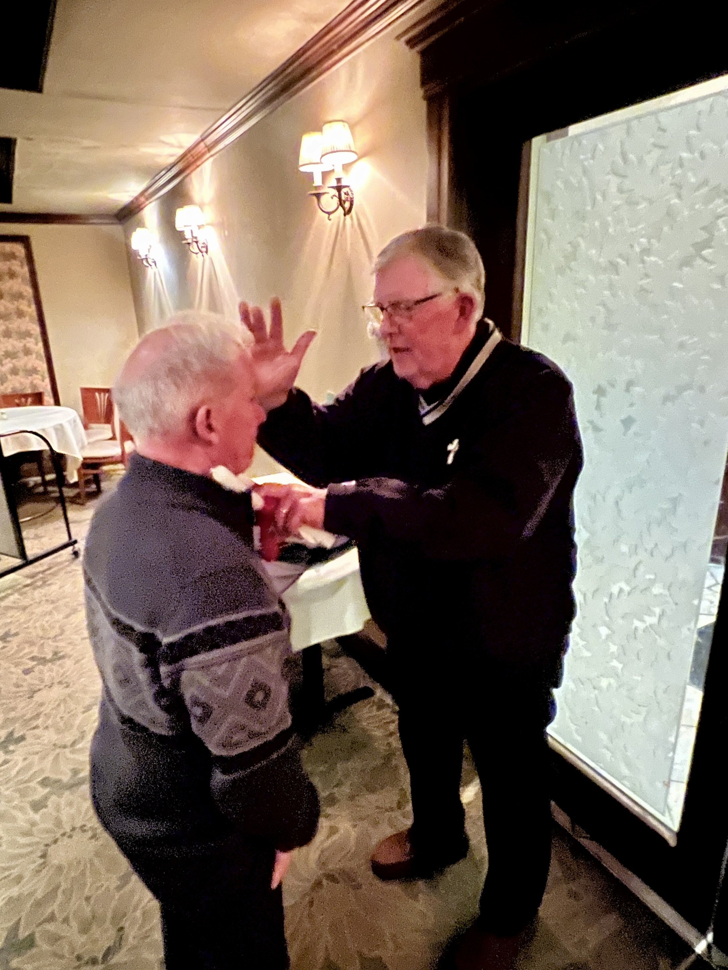 Two elderly men interacting, one placing his hand on the other's forehead, in a dimly lit room with decorative lighting and carpet.