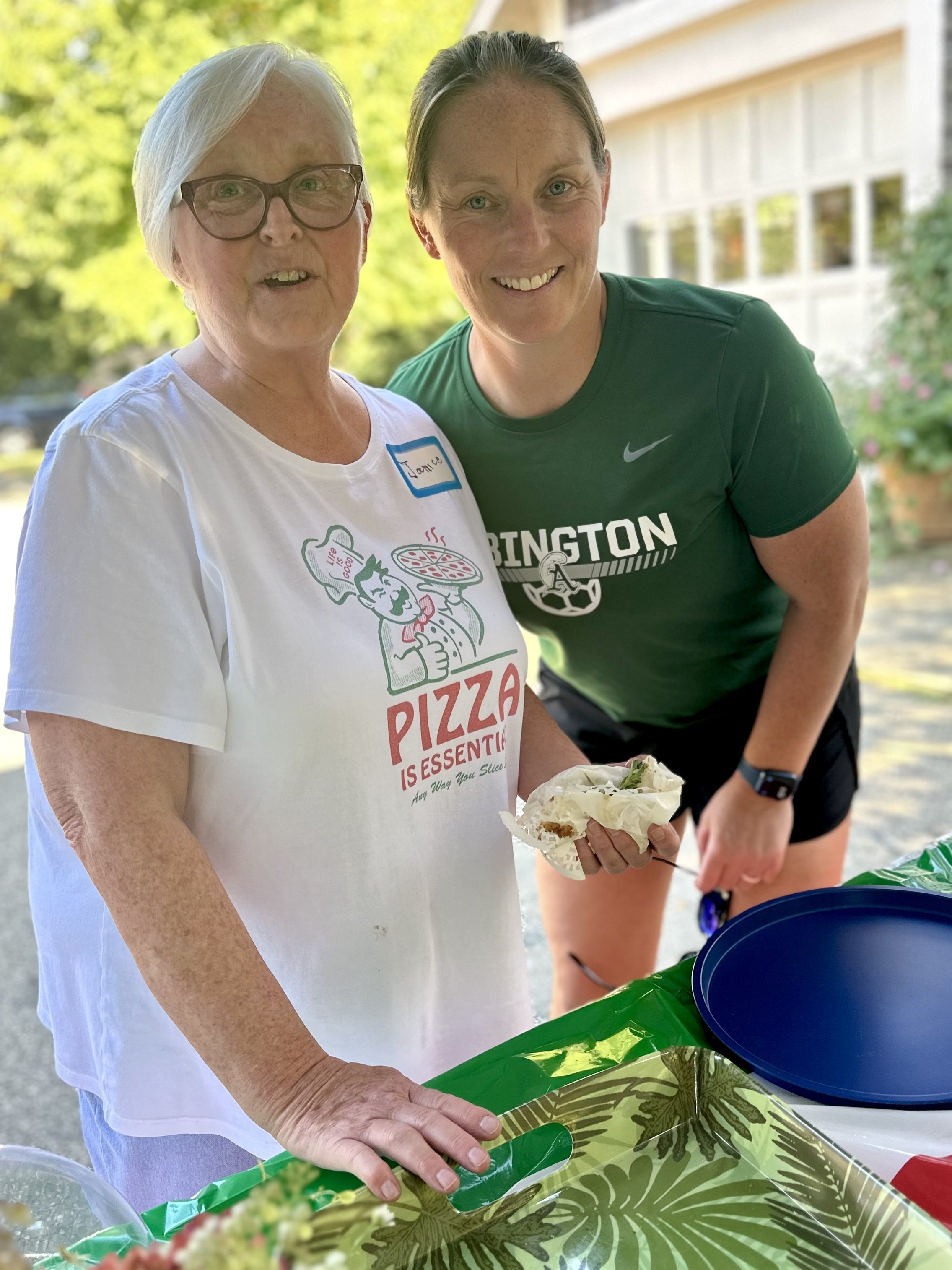Two people smiling outdoors at a table. One wears a white shirt with a pizza graphic, and the other wears a green sports shirt. They appear to be at an informal gathering or picnic, with a covered table in front of them and a blue plate visible.