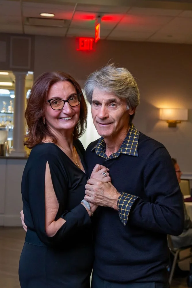 A smiling woman and a man are dancing closely together indoors, holding hands. The woman has shoulder-length brown hair and glasses, and is wearing a black dress with open sleeves. The man has gray hair and is wearing a black sweater with a yellow ch