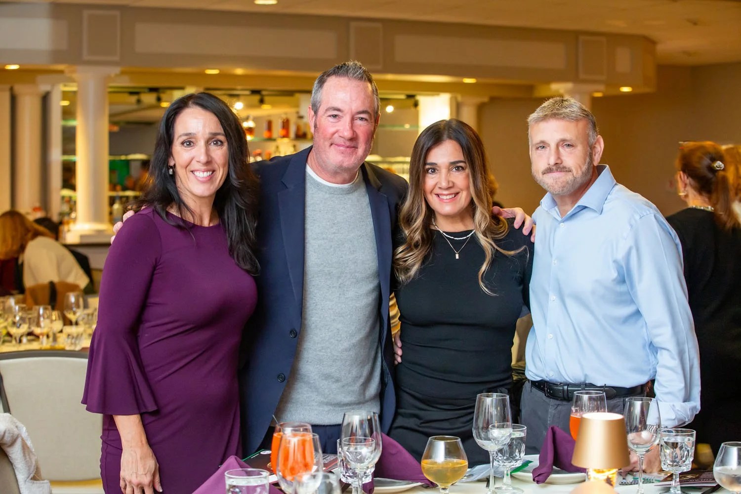 Four people standing together at a dinner event, smiling at the camera, with a table set with wine glasses and purple napkins in the foreground.