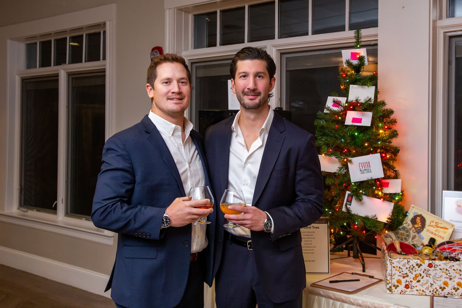 Two men in business suits holding drinks and smiling at an indoor holiday party, with a decorated Christmas tree and holiday-themed decorations in the background.