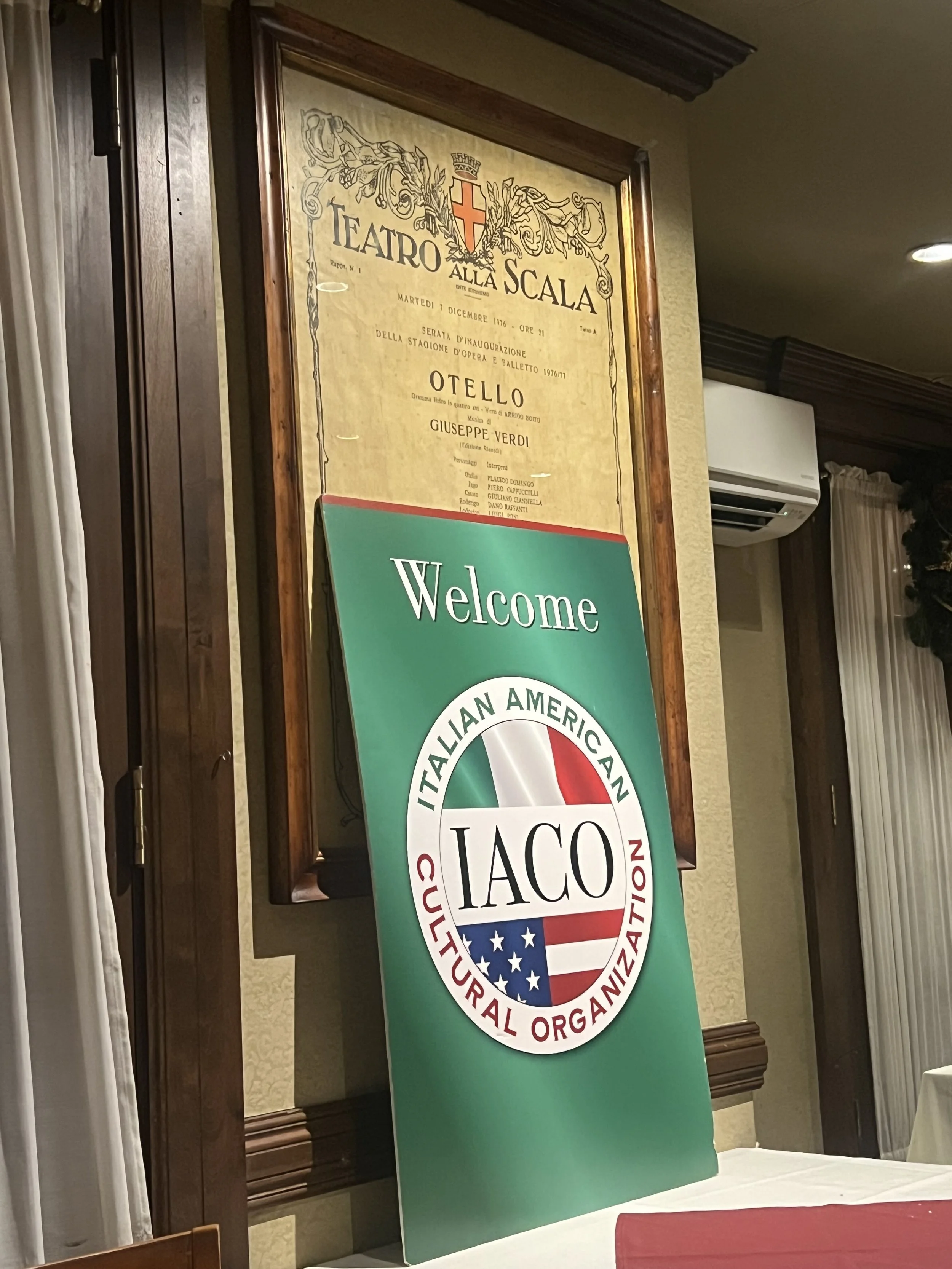 A welcome sign for the Italian American Cultural Organization (IACO) placed in front of a poster for Teatro alla Scala at an indoor event.